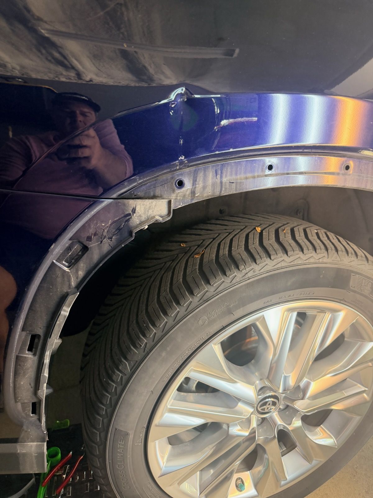 Blue car front wheel with chrome rim and tire under a fender arch, with a person reflected nearby