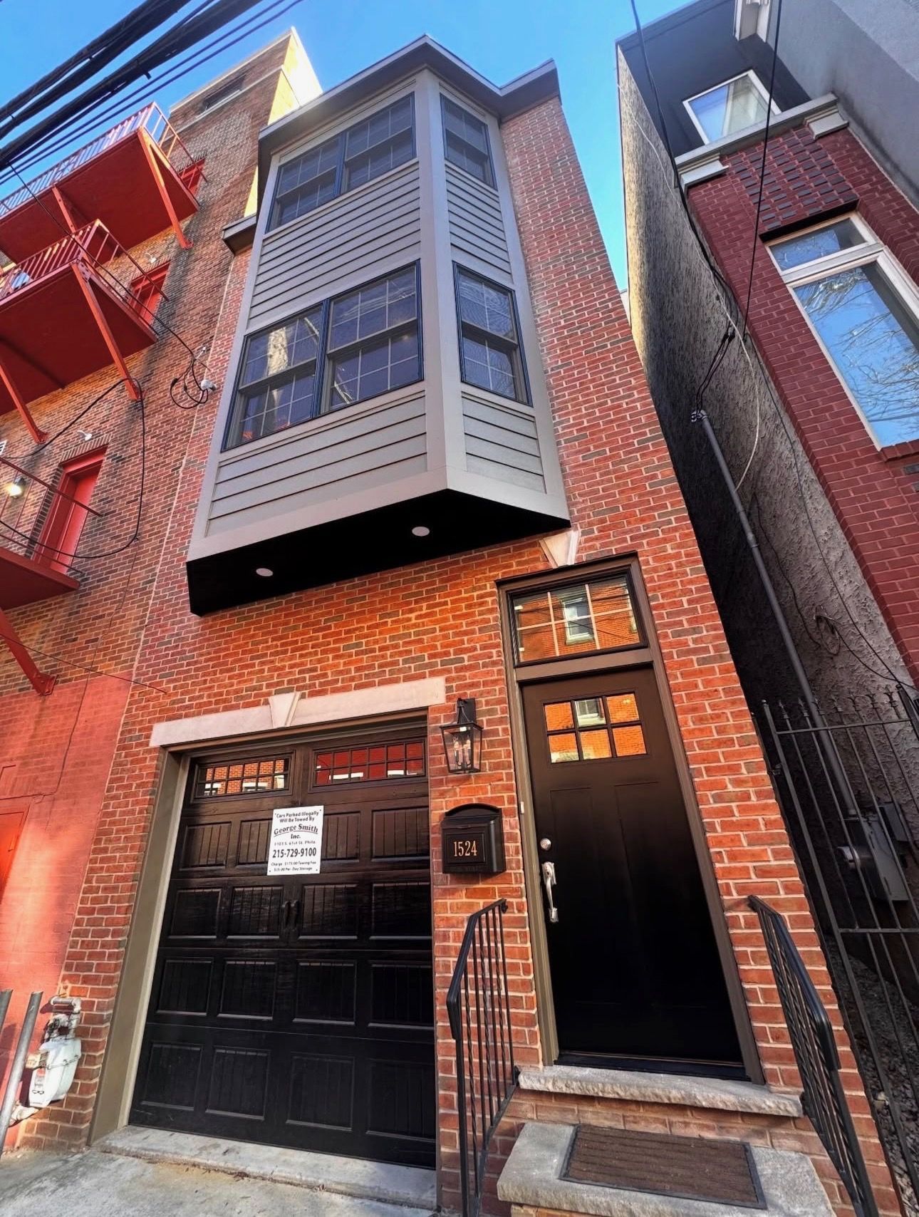 Narrow brick townhouse façade with black garage door and bay window above, viewed from a steep alley.