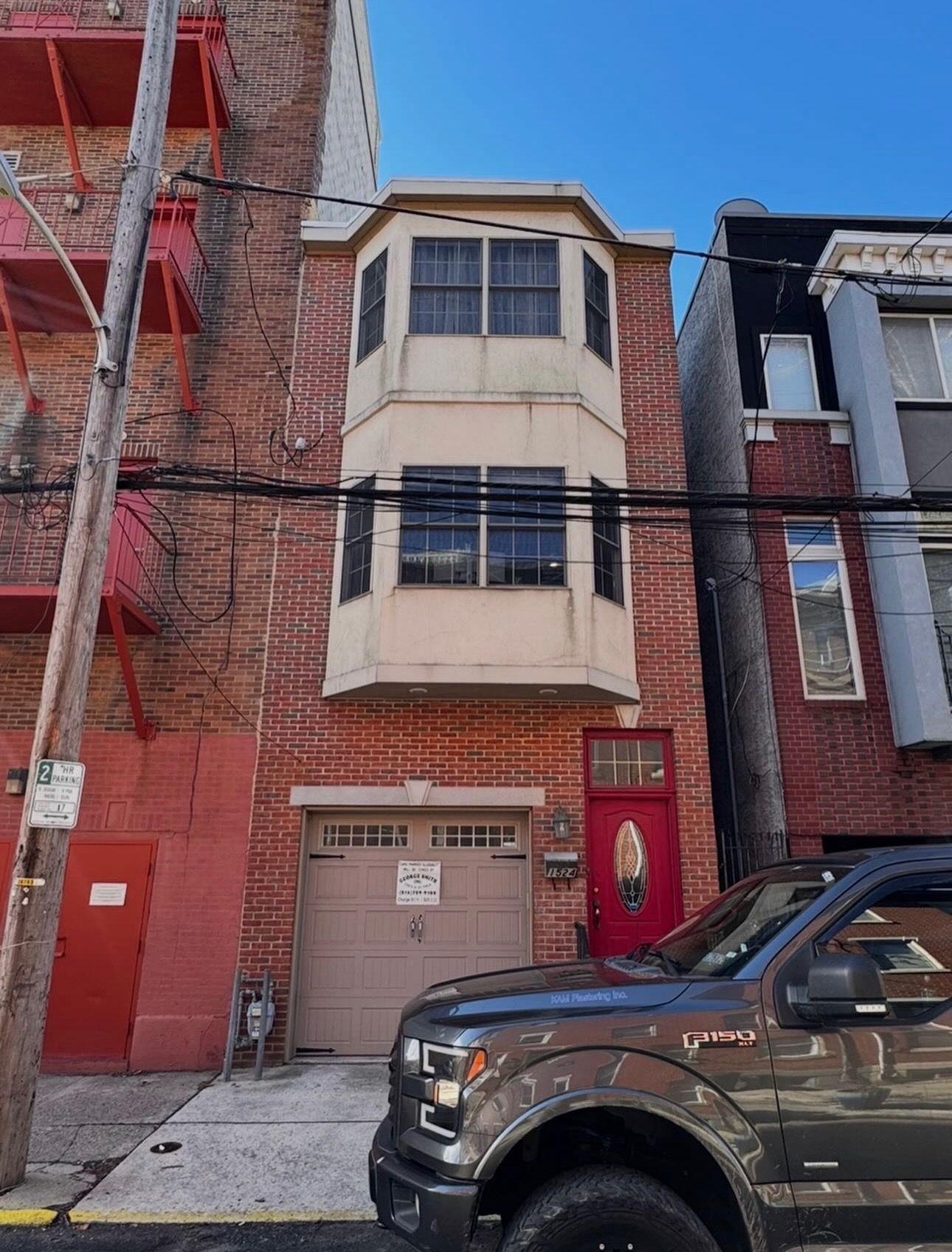 Narrow brick row house with bay windows and red door on a city street, with a black pickup truck parked in front