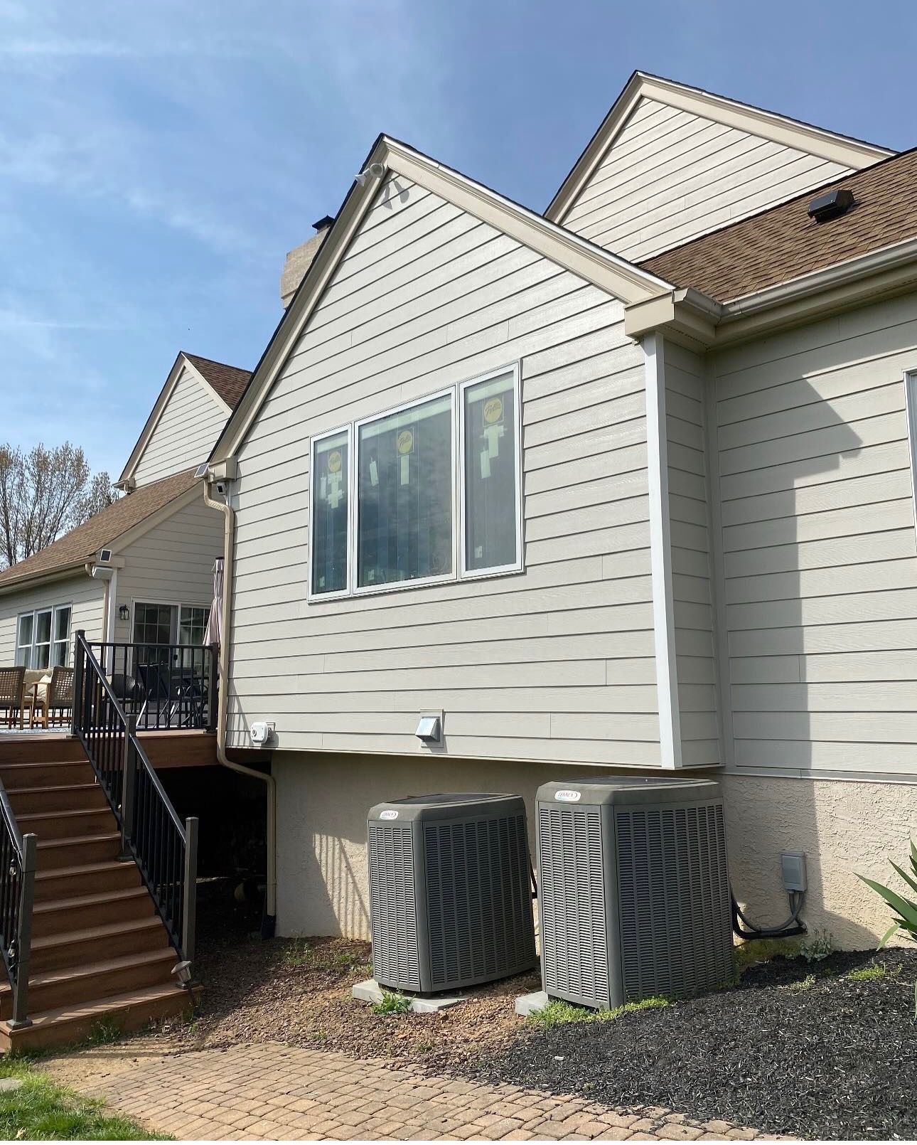 Exterior of a beige house with a large window, stairs, and two HVAC units on a gravel yard