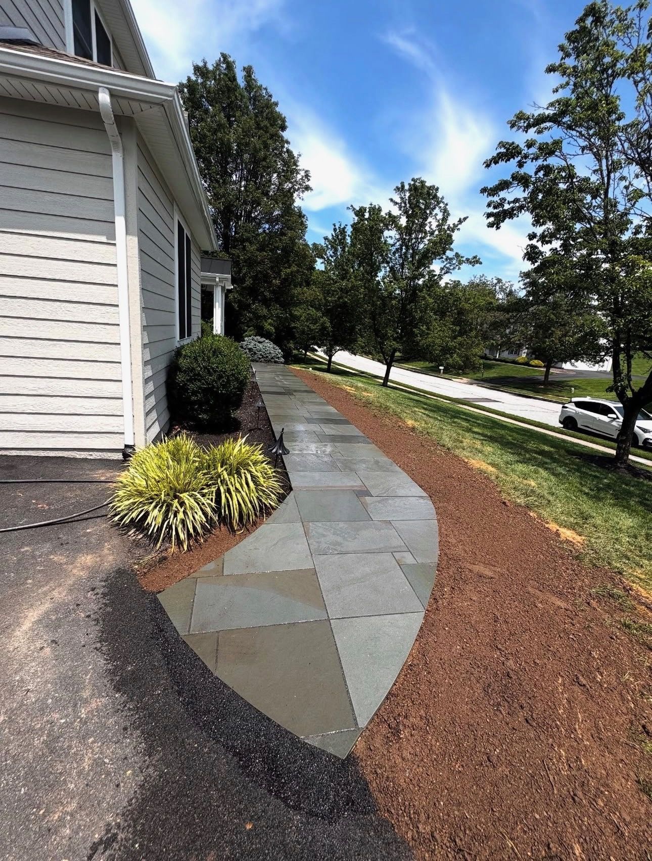 Stone walkway beside a house, bordered by mulch, shrubs, and trees under a bright sky.