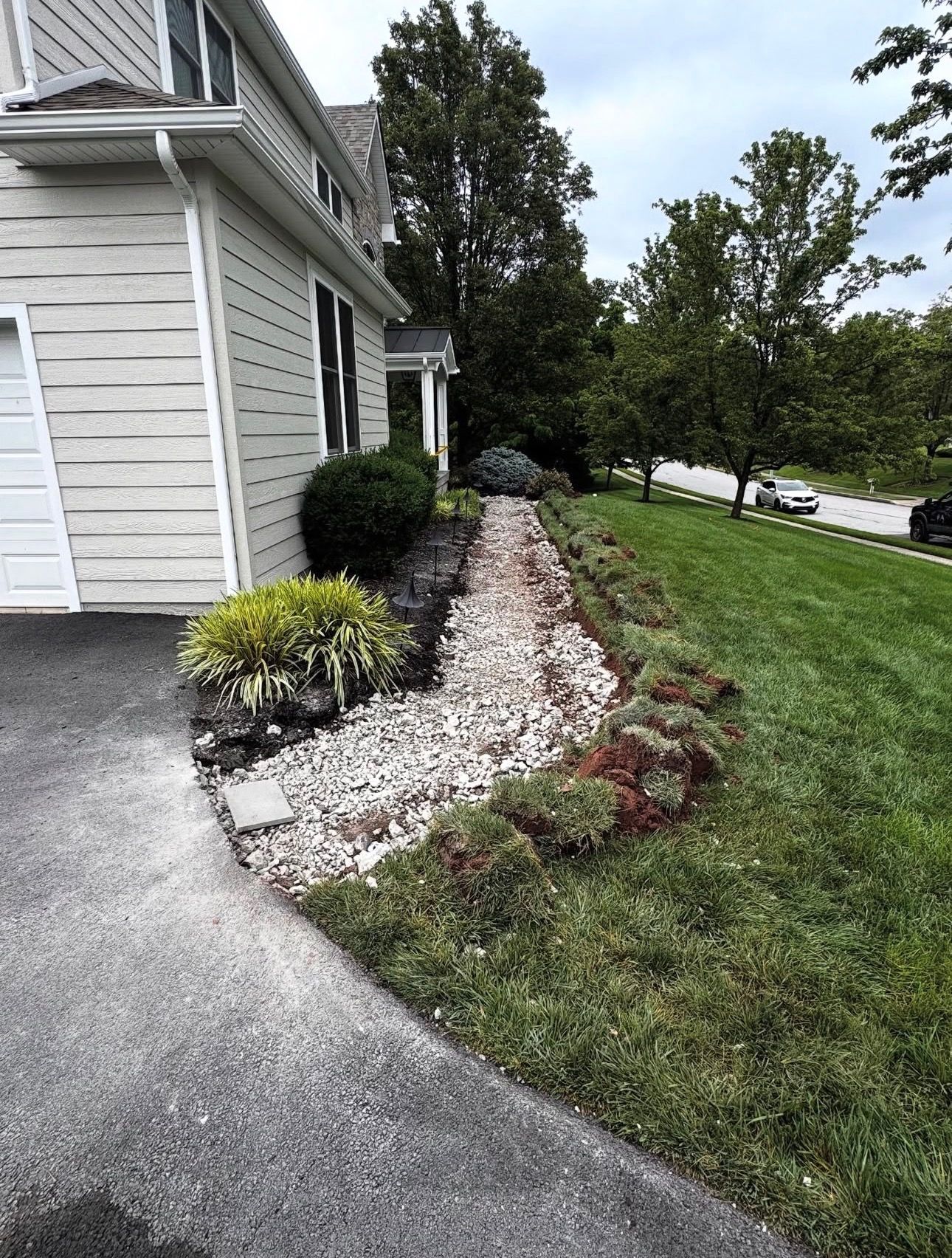 House side yard with white gravel border, shrubs, and a grassy slope beside a driveway and street.