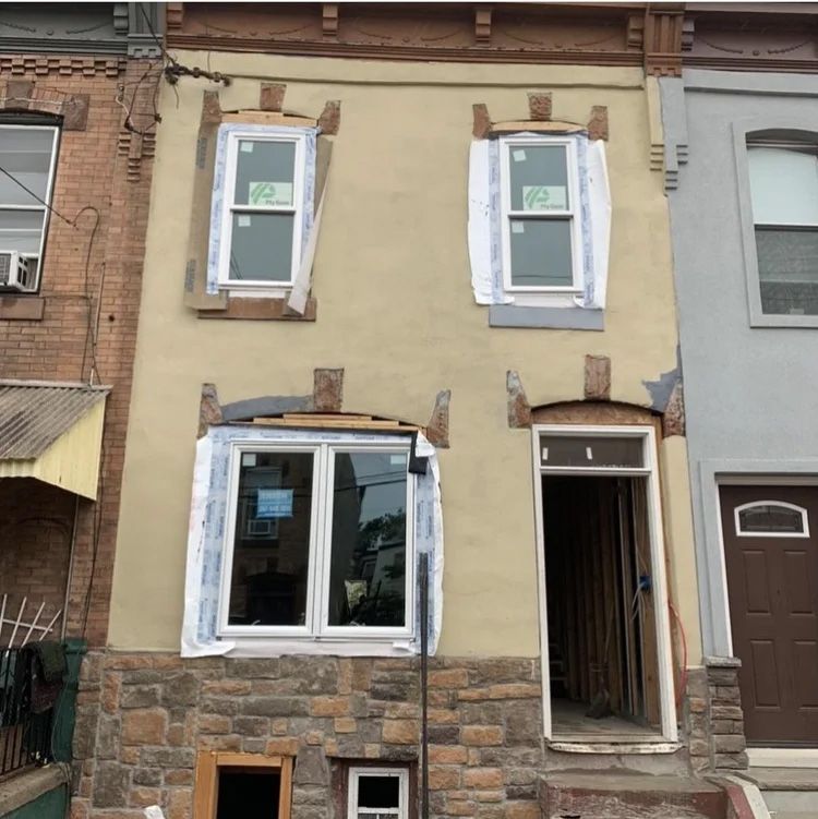 Exterior of a two-story rowhouse under renovation, with new windows and exposed stone at the base.