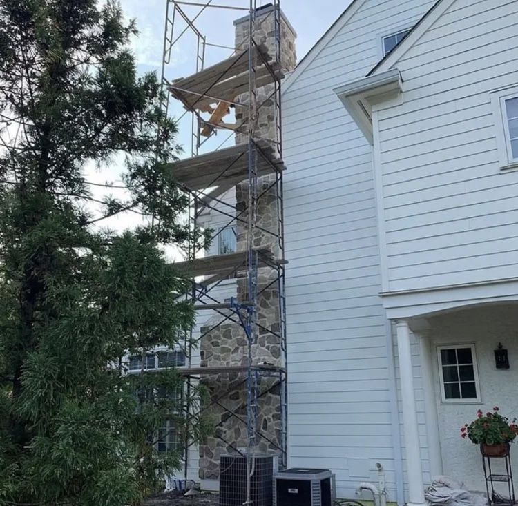 Scaffolding surrounds a stone chimney on a white-sided house for repairs.