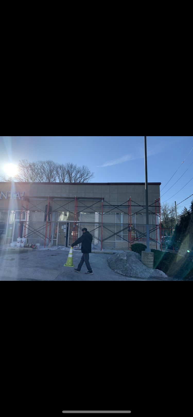 Person walks in front of a building under construction, carrying a bright yellow item. Bright sun.