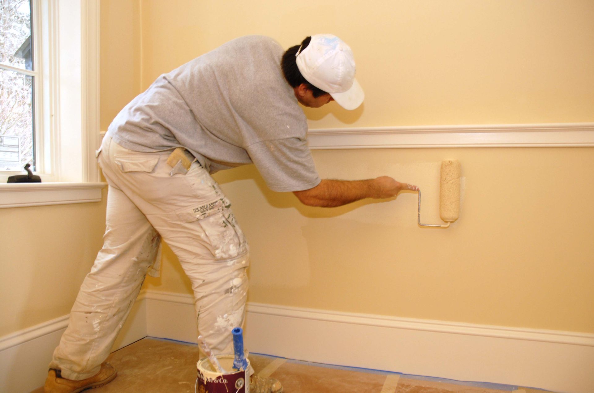 Person painting a beige wall with a roller in a room with wood floors and a window