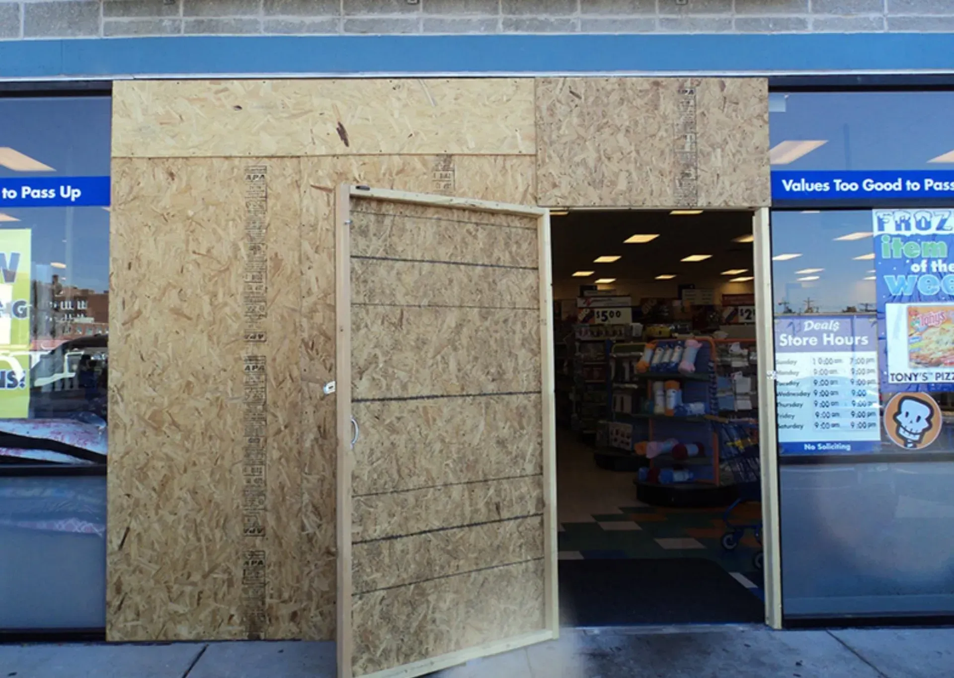 A store entrance boarded up with plywood, doorway ajar, interior visible, reflecting sunlight.