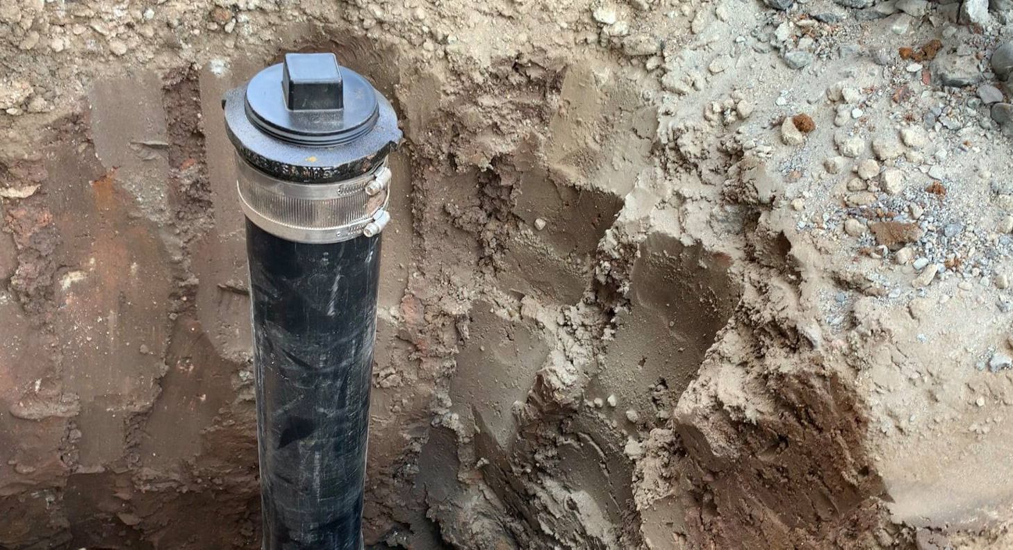 A black vertical pipe with a capped, silver-banded end stands in a small, excavated hole in the dirt.