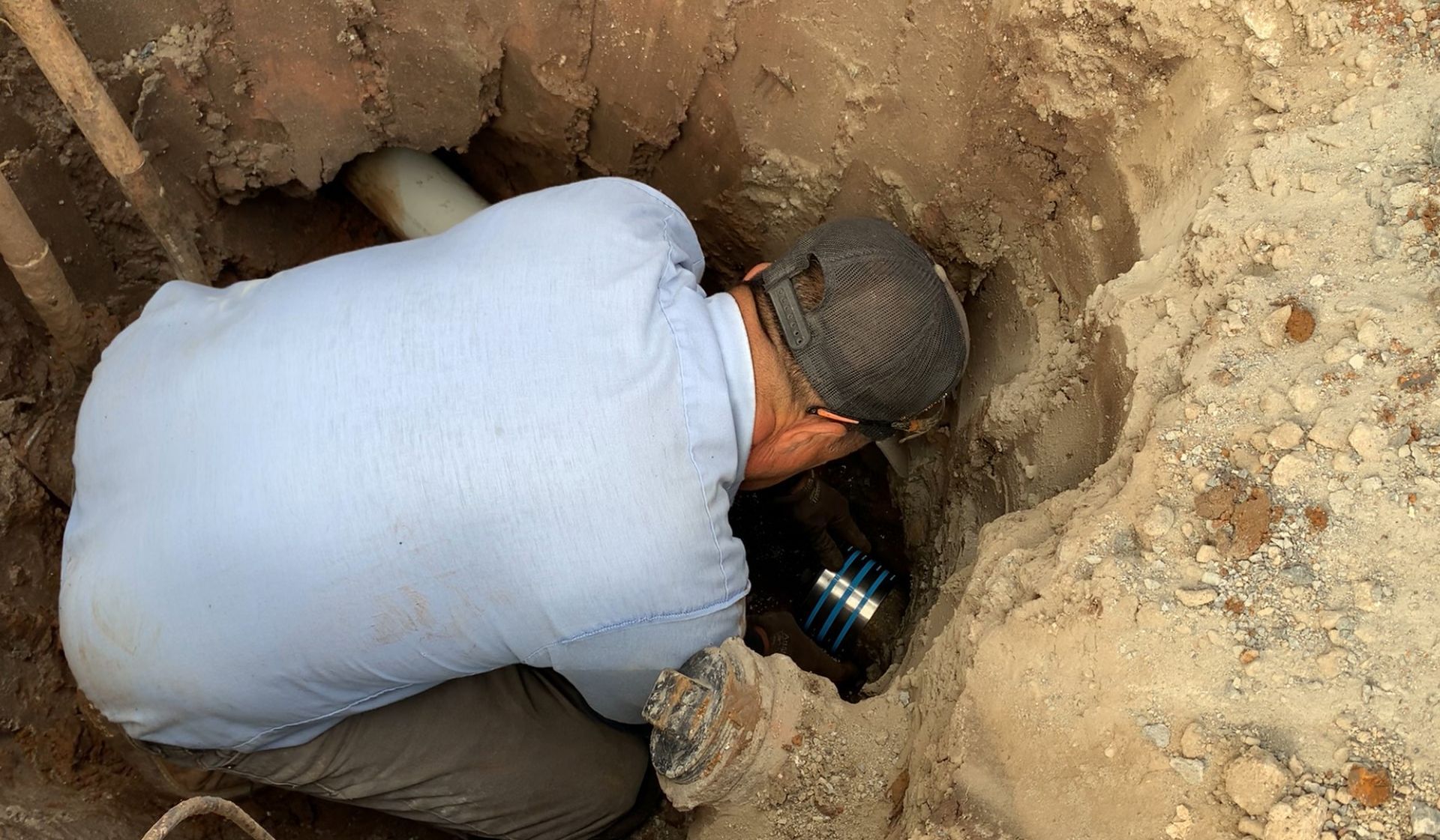 A person in a light blue shirt and dark cap kneels in a dirt trench, working on pipes or electrical components underground.