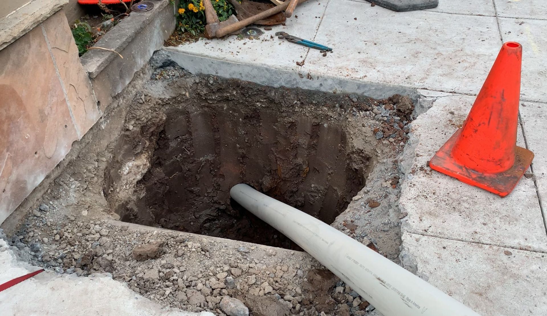 A square excavation hole in a concrete patio with a single white PVC pipe extending into the dark earth.