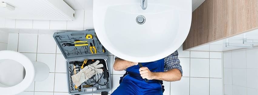 A plumber in blue overalls works under a bathroom sink, with a toolbox nearby on a white tiled floor.