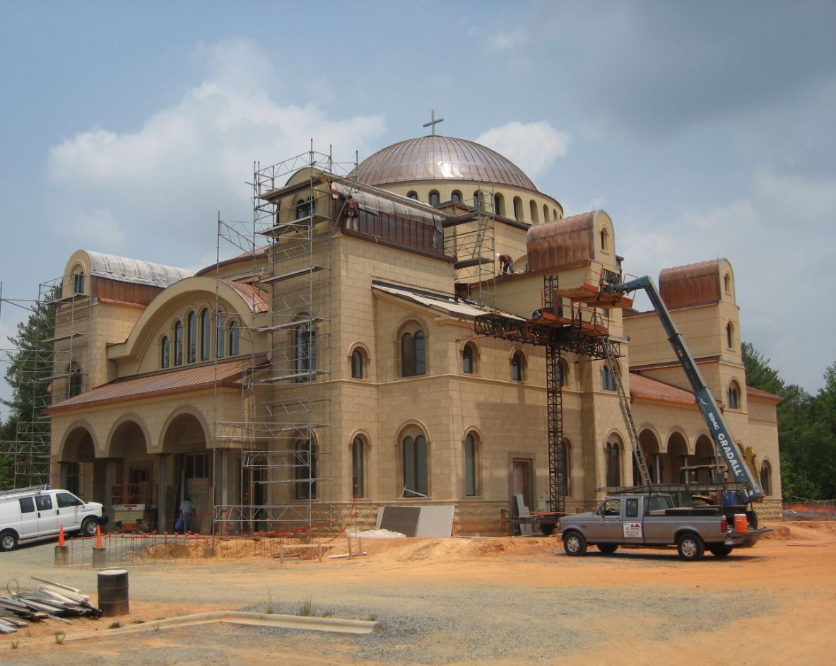 Church under construction with dome, scaffolding, and cherry picker in a dirt lot.
