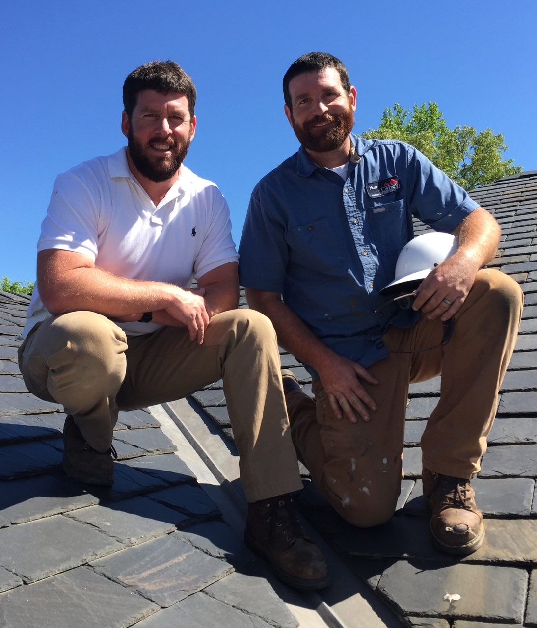 Two men on a slate roof, both with beards, wearing work clothes. One holds a white helmet. Blue sky background.