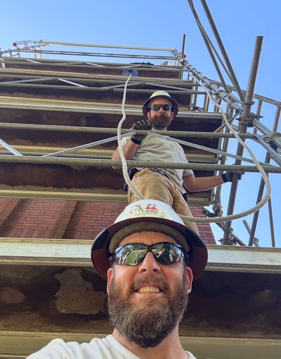 Two construction workers on scaffolding, one taking a selfie, both wearing hard hats and sunglasses. Brick building in the background.