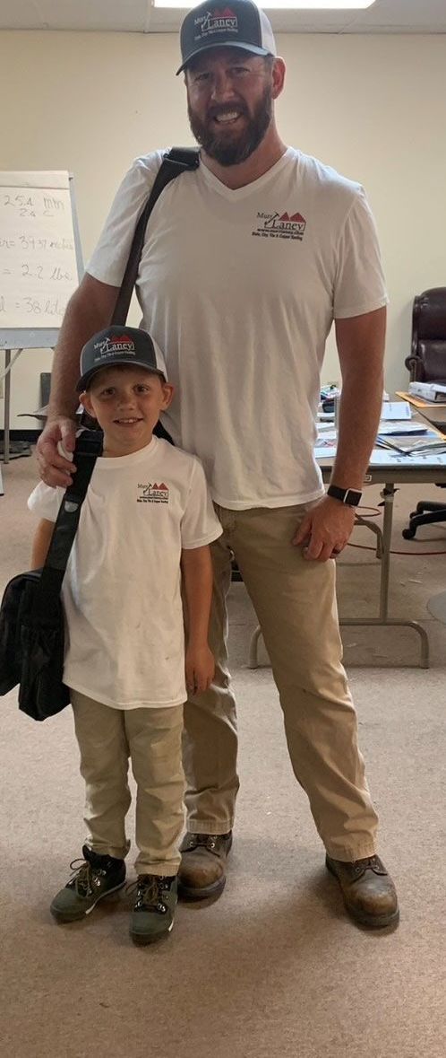 A man and a young boy pose together indoors. Both are wearing white t-shirts, baseball caps, and khaki pants. The boy holds a black bag over his shoulder.