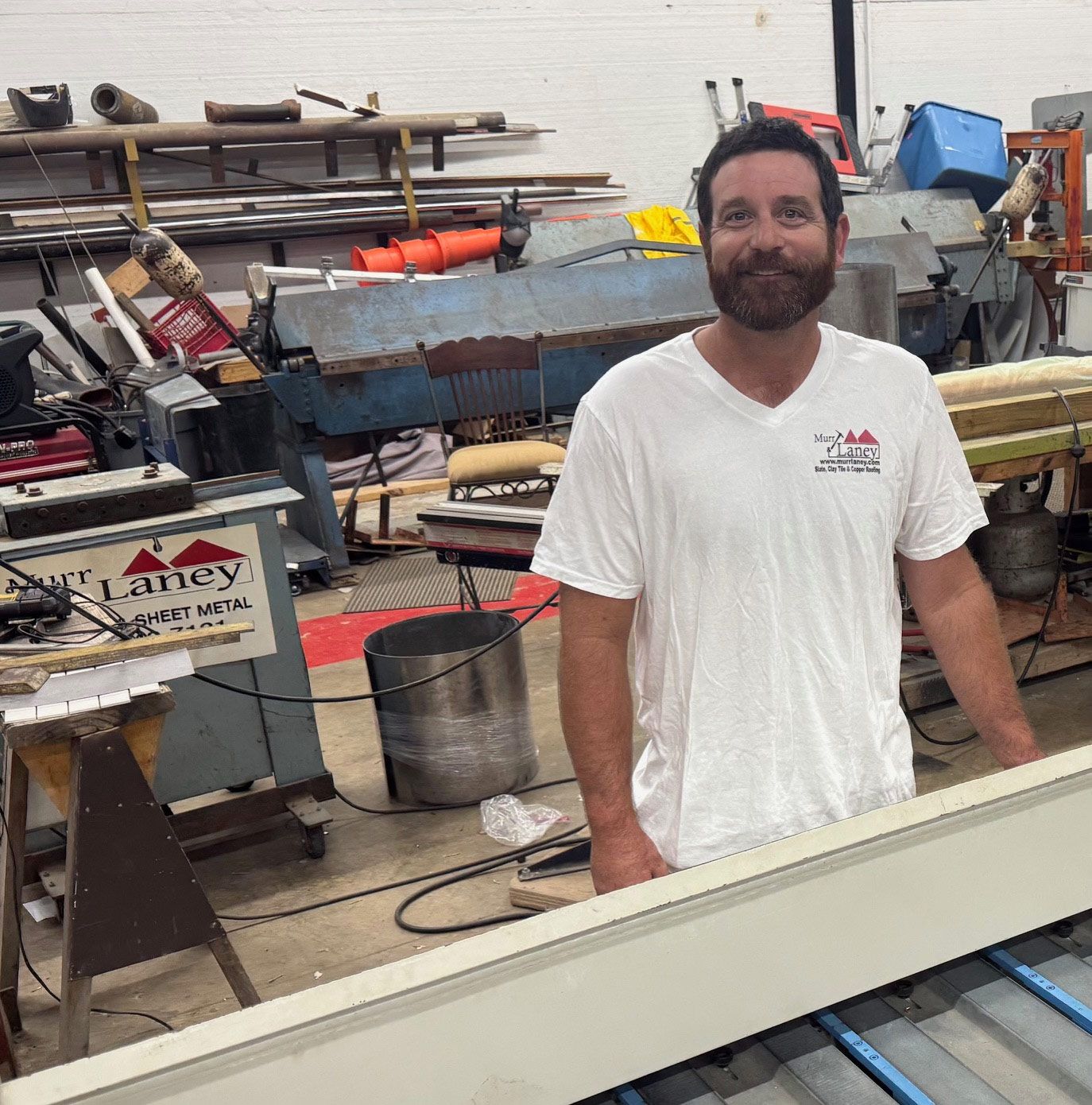 A man with a beard in a white shirt smiles in a workshop. He stands next to a piece of trim.