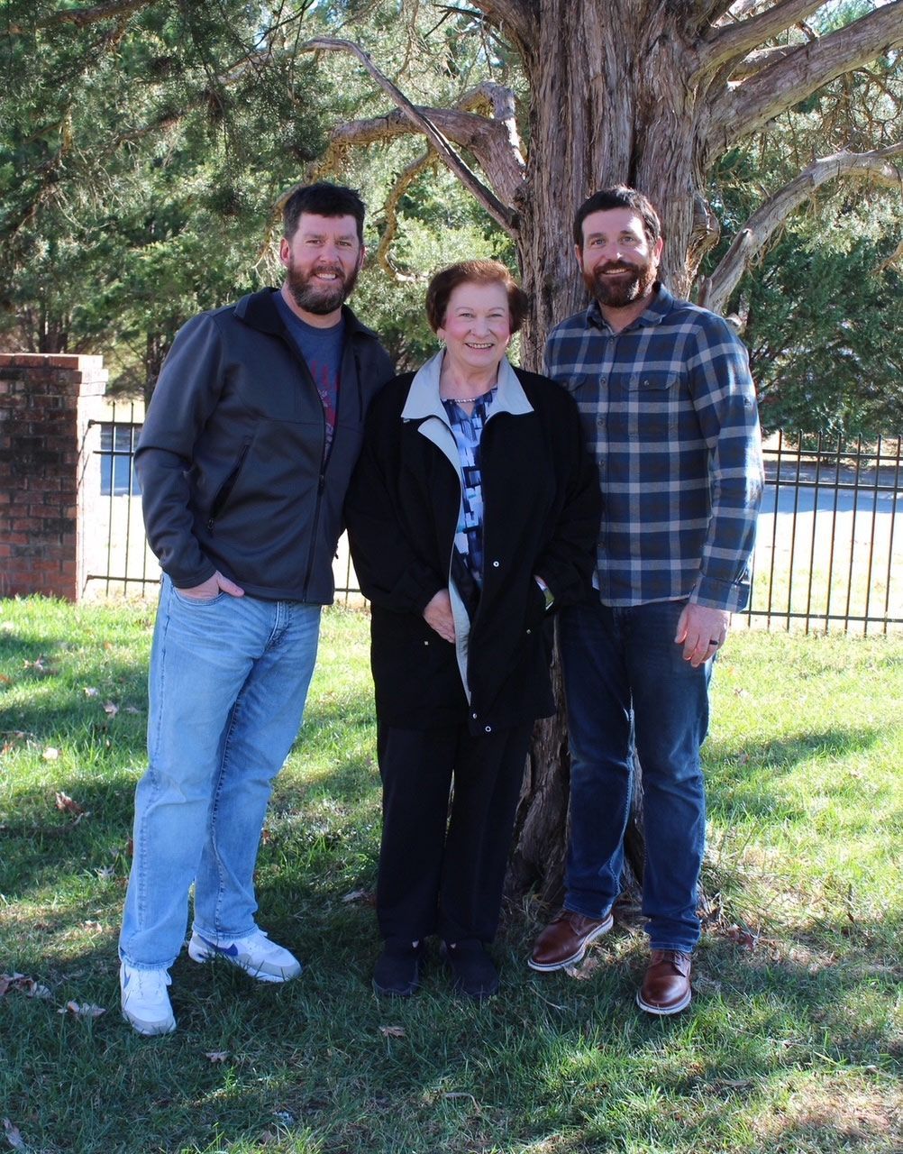 Three people standing by a tree outdoors. Woman in black jacket between two men with beards and casual clothes.