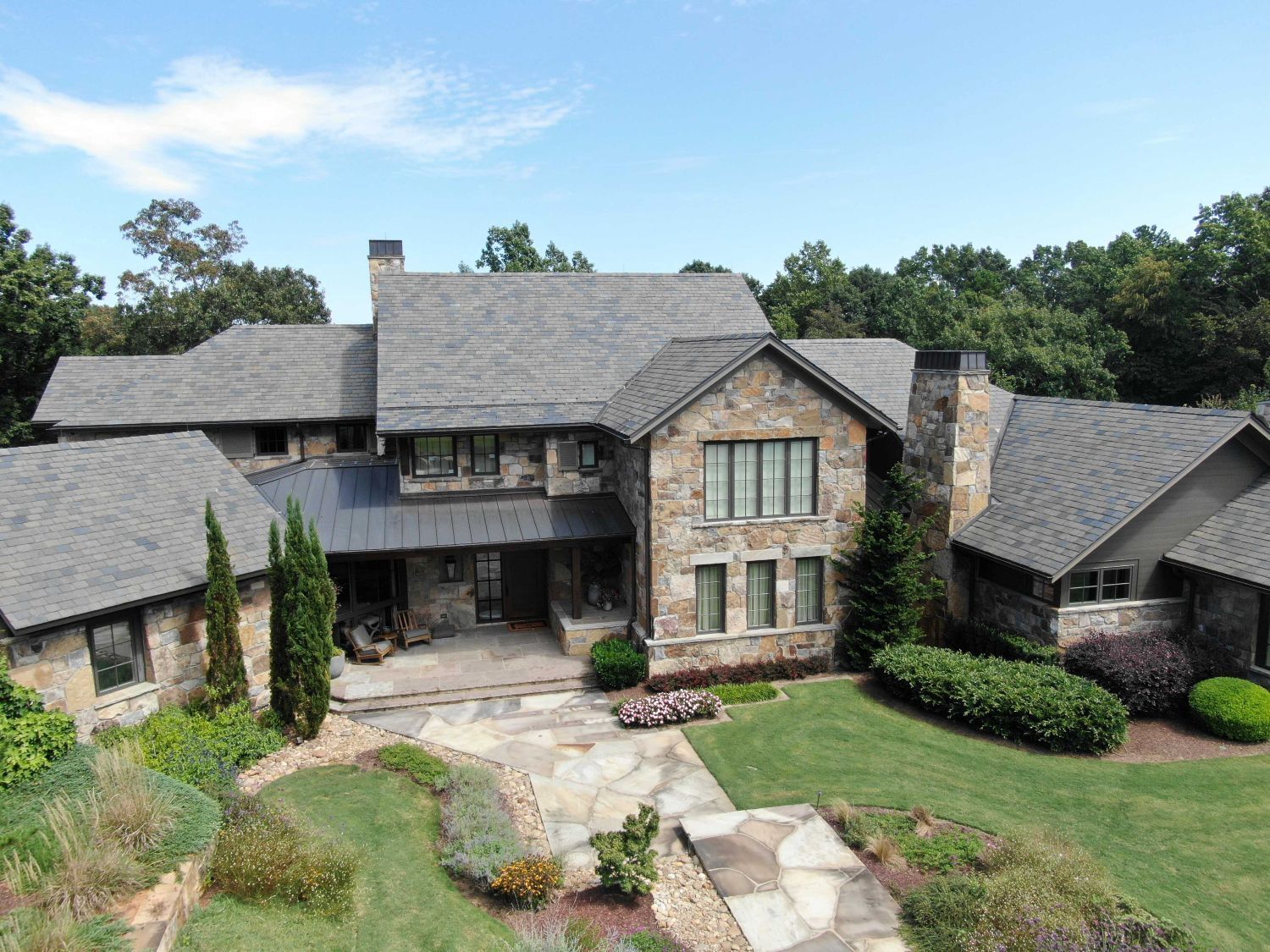 Stone house with gray roof, green lawn, and a stone pathway. Trees surround the home under a blue sky.