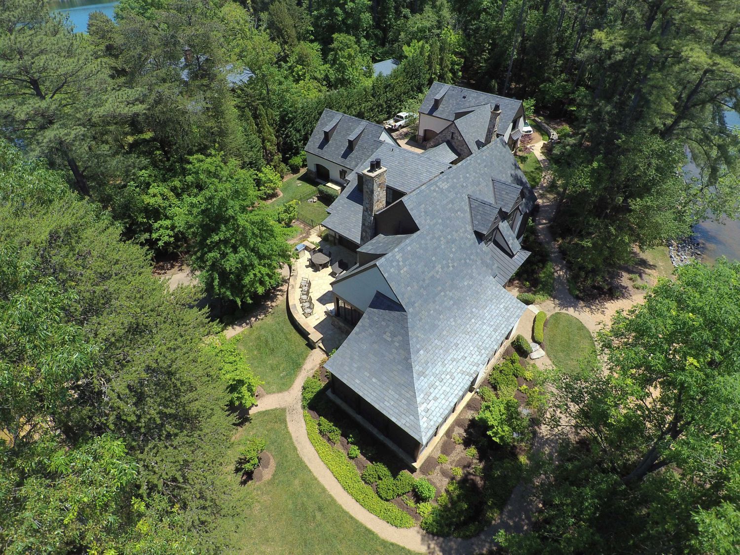 Aerial view of a large house with a dark gray roof surrounded by lush green trees and a lake.