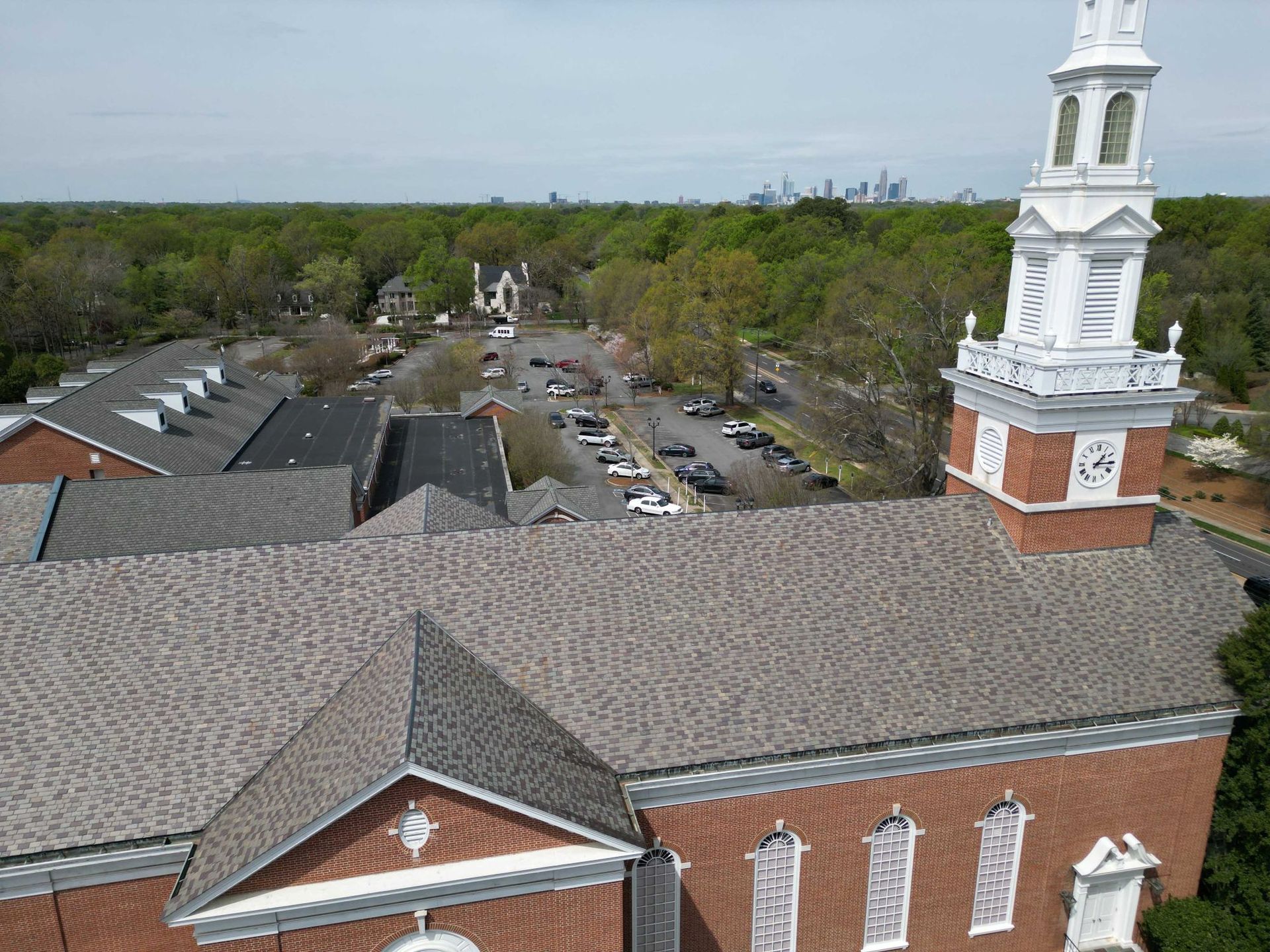 Aerial view of a brick church with a white steeple, surrounded by green trees and buildings. Cars are parked in the background.