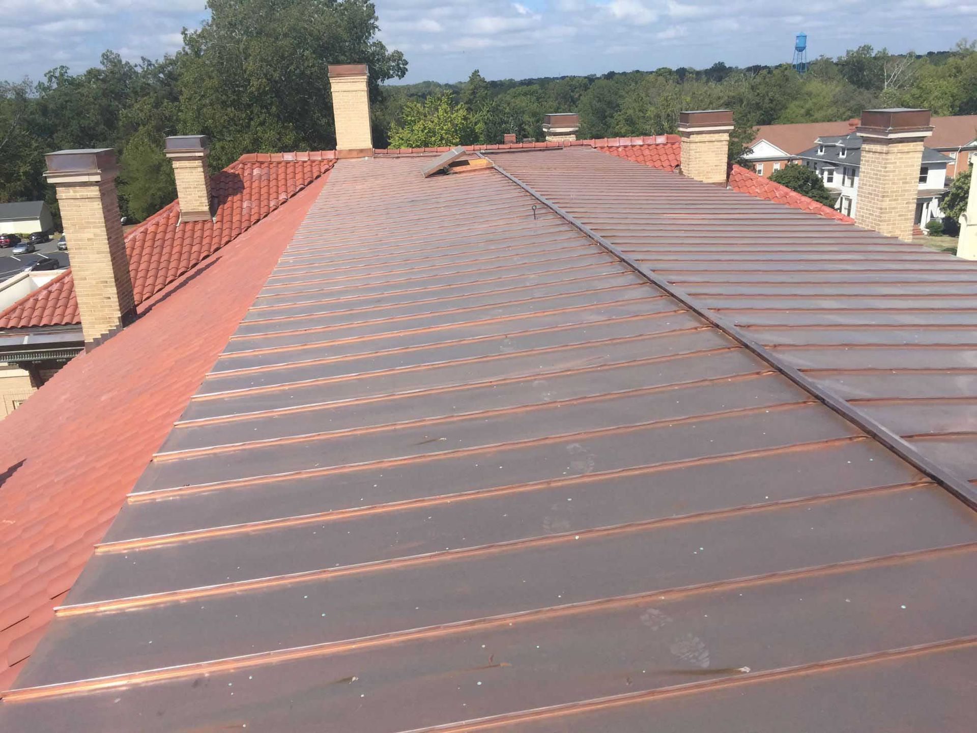 Copper metal roof with vertical seams. Several brick chimneys and trees in the background under a blue sky.