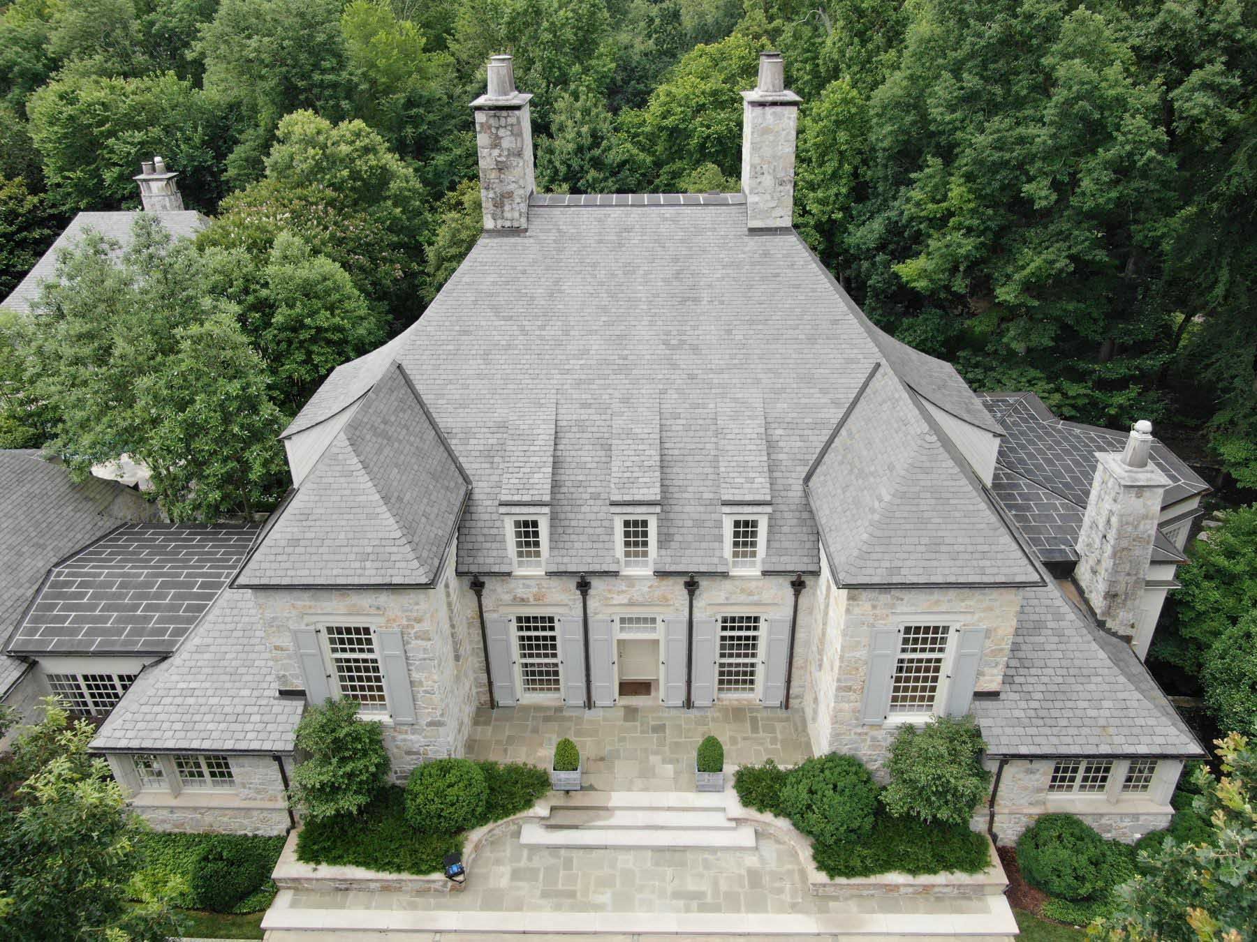 An aerial view of a large stone house with a gray shingled roof and a manicured front yard surrounded by trees.