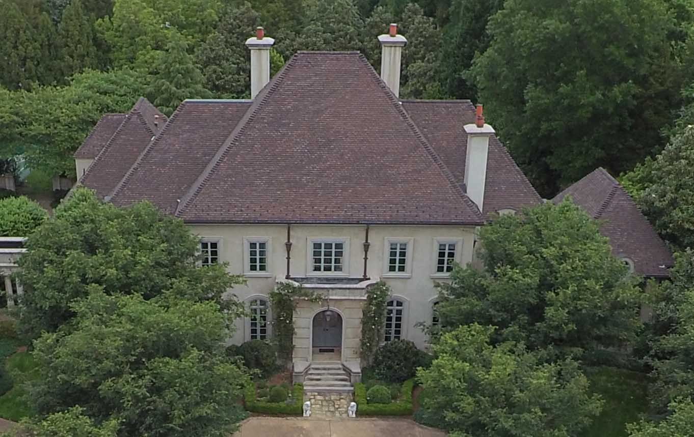 A large, light-colored mansion with a brown tiled roof is surrounded by green trees.