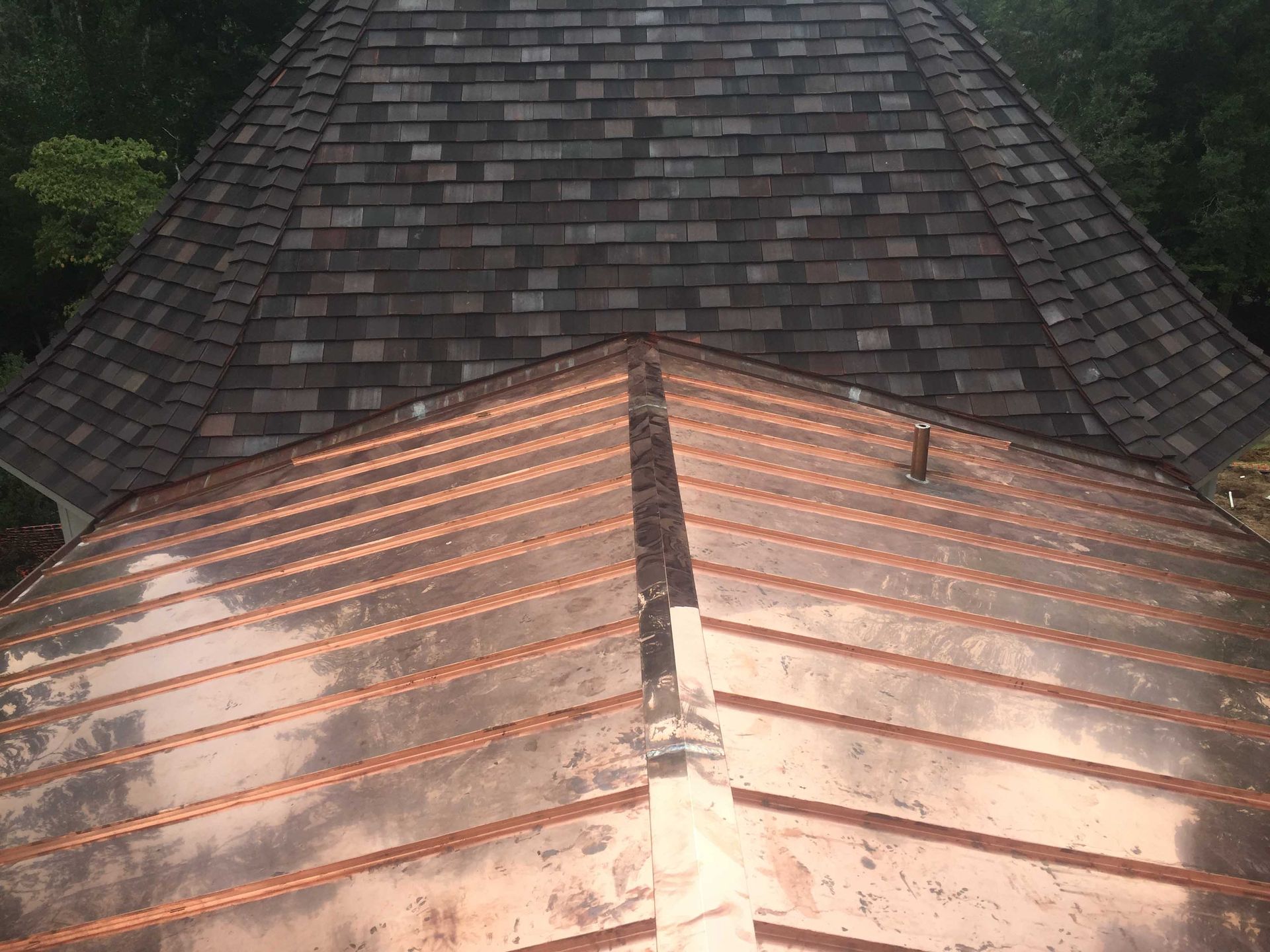 A top-down view of a copper roof in the foreground, with a weathered, shingled roof in the background.