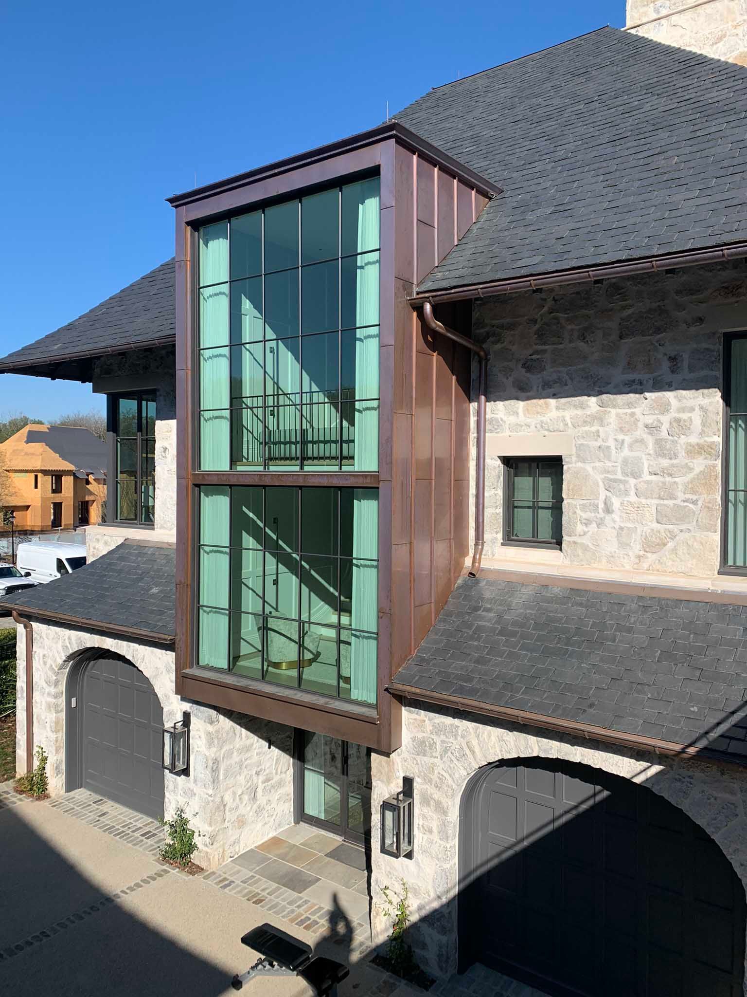 A modern building with a copper-clad glass staircase tower. Stone exterior, dark gray roof, and arched garage doors.