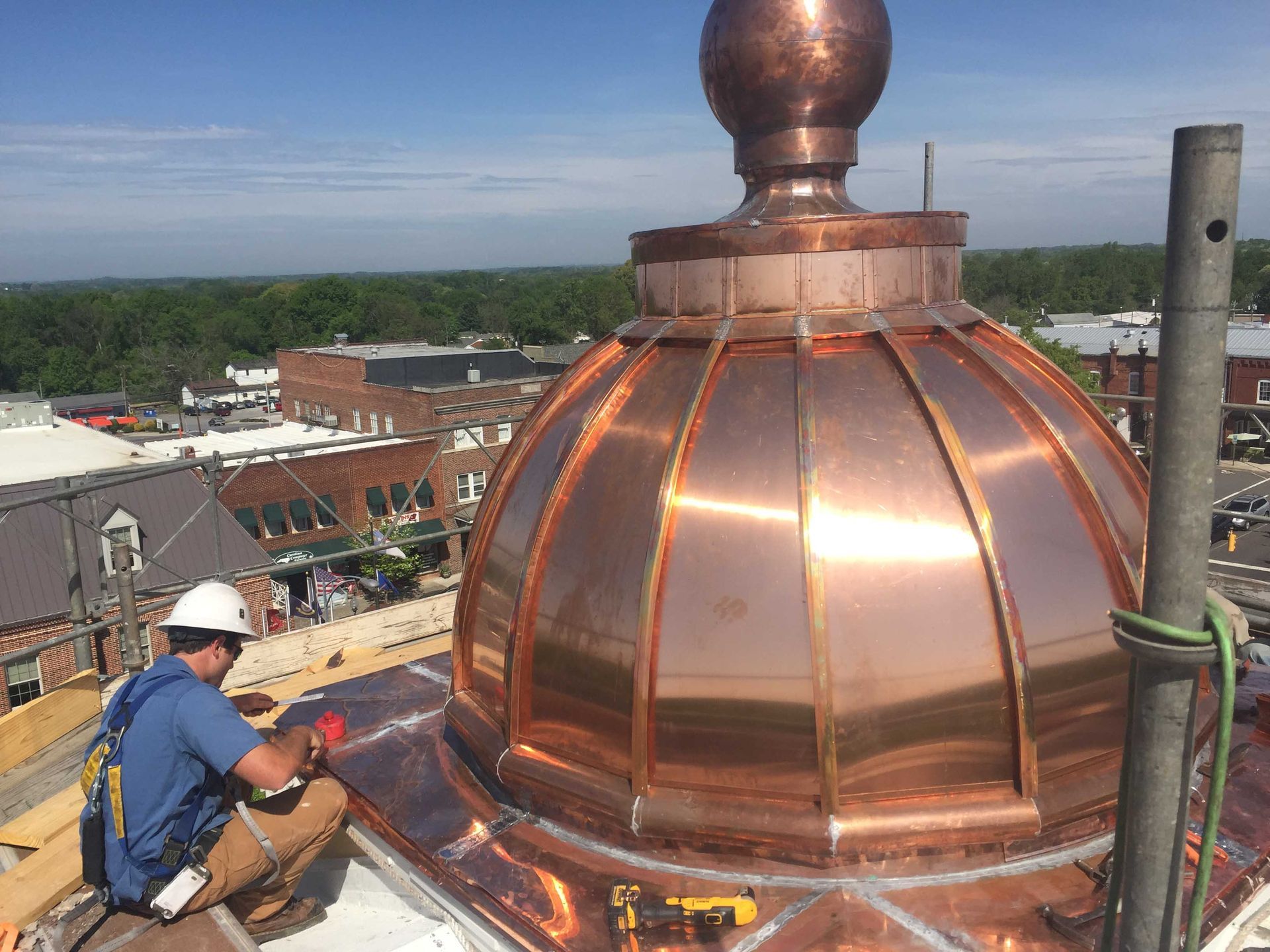 A worker installs copper sheeting on a dome-shaped roof with a cityscape in the background. The dome is shiny copper with a bronze finial.