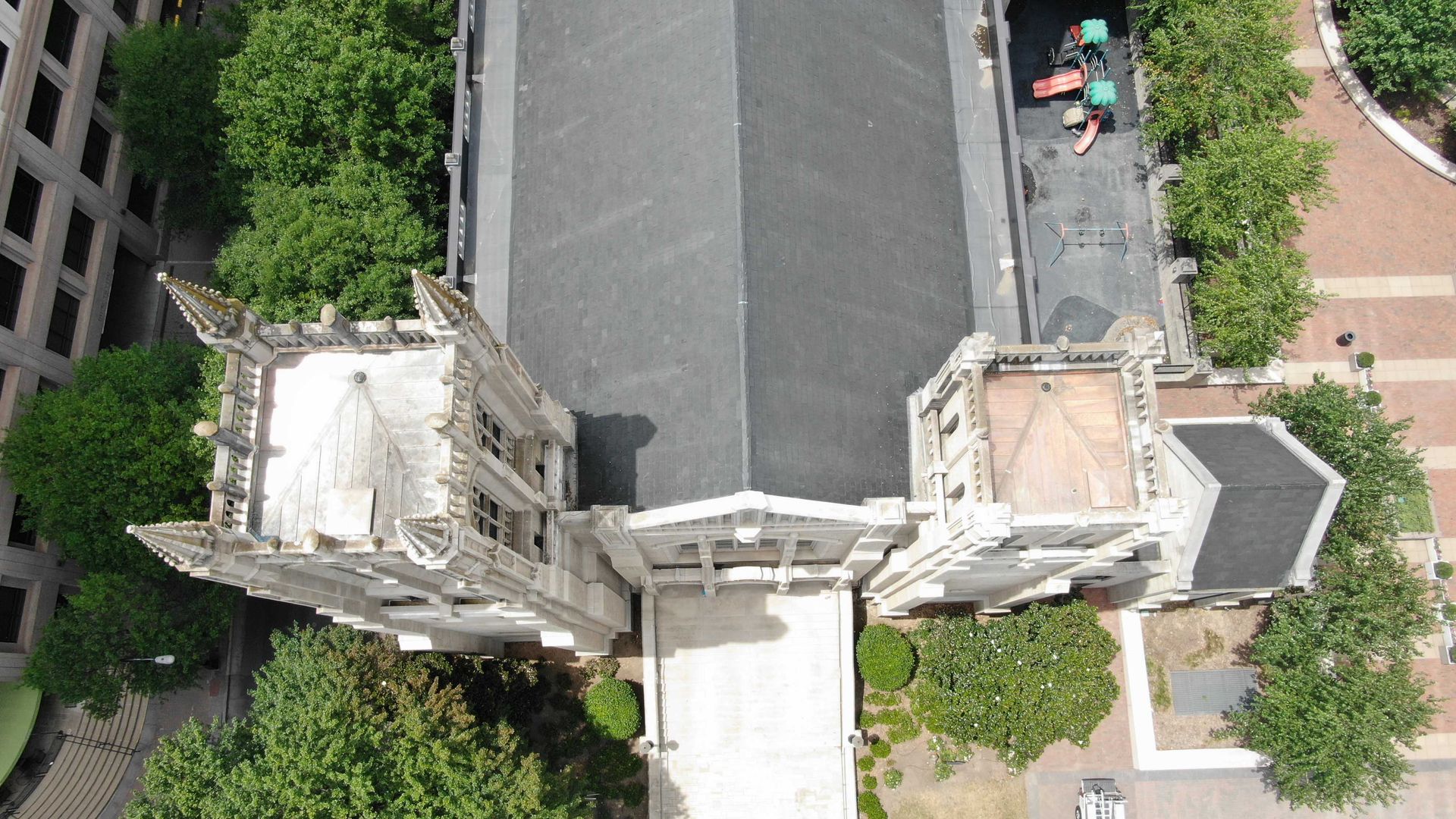 Overhead view of a gray stone building with dark roof and surrounding green trees and paved areas.