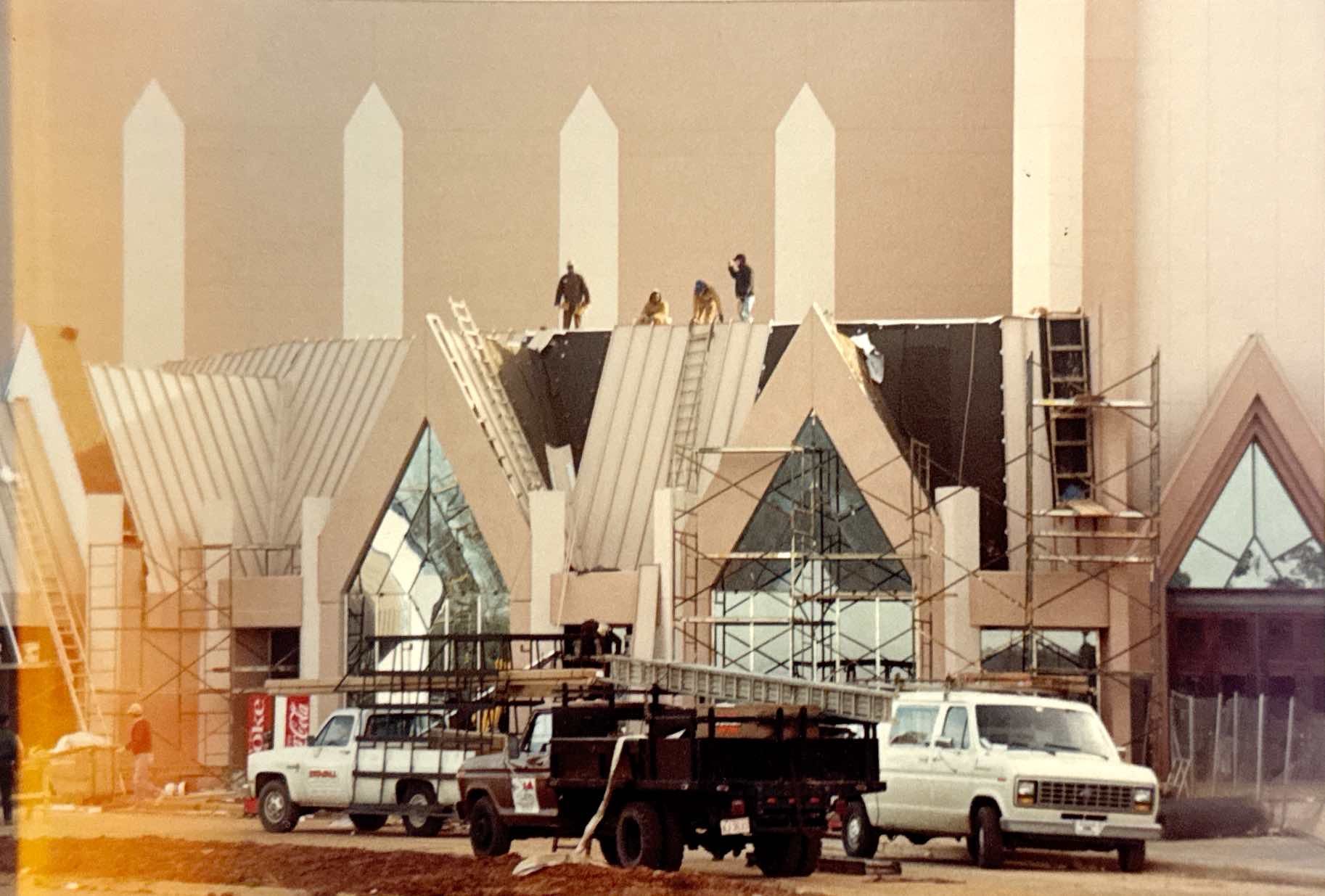 Construction of a light-colored building with pointed roof sections and large triangular windows. Workers are visible on the roof.