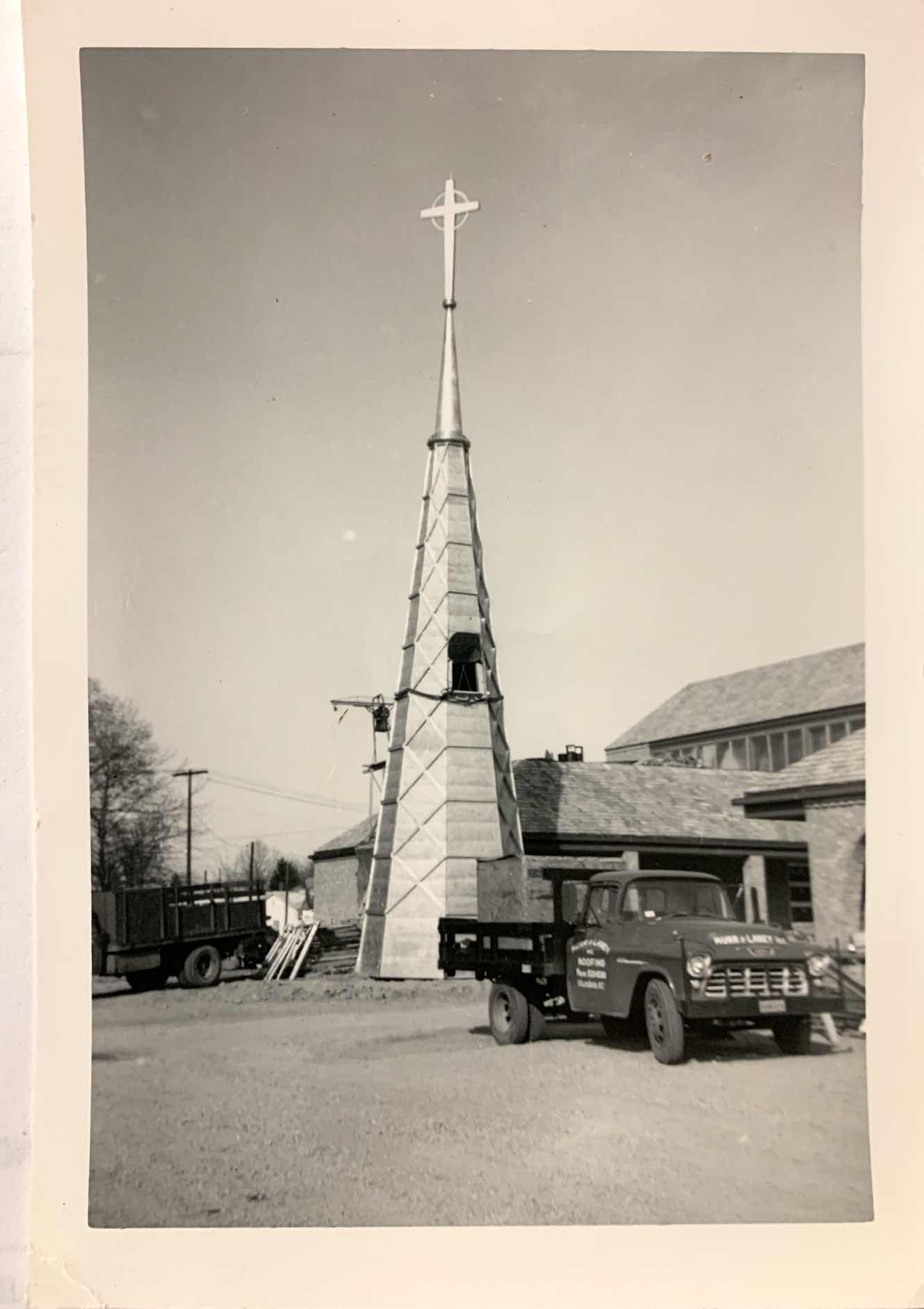 Black and white photo of a church steeple under construction, with a cross on top, surrounded by scaffolding and trucks.