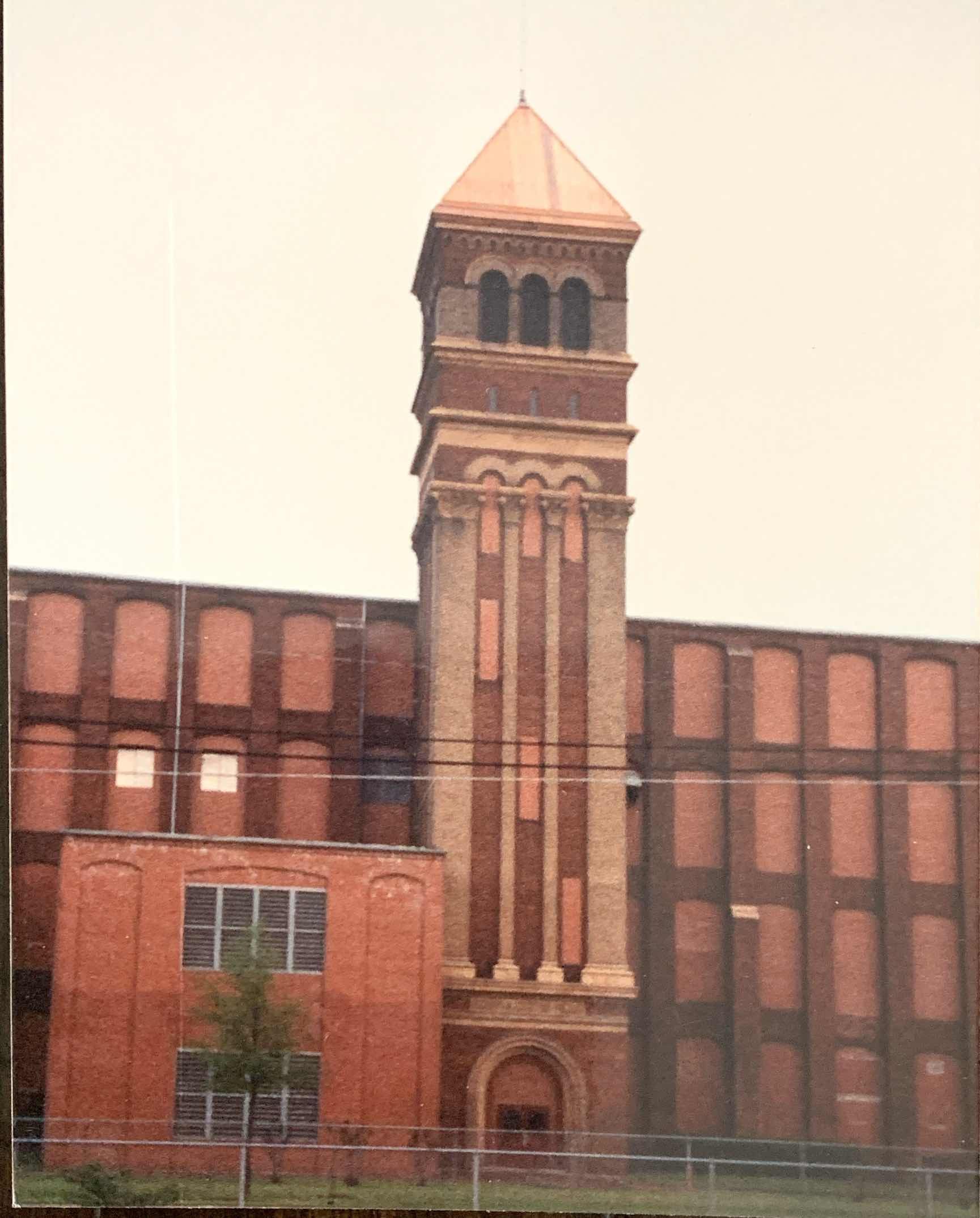 Tall brick factory building with a prominent clock tower. The tower features a copper-colored roof and arched windows.