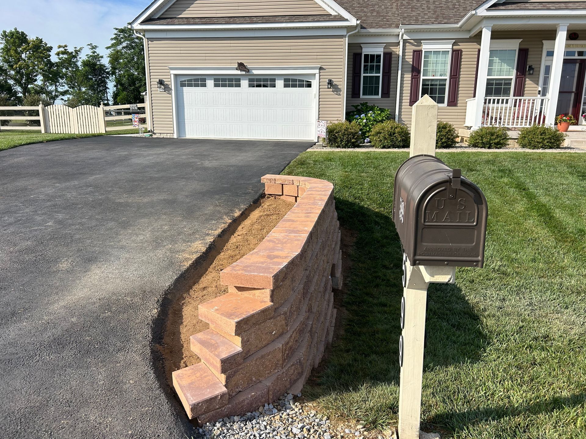 A curved stone retaining wall borders a dark asphalt driveway next to a grass lawn and a mailbox in front of a house.