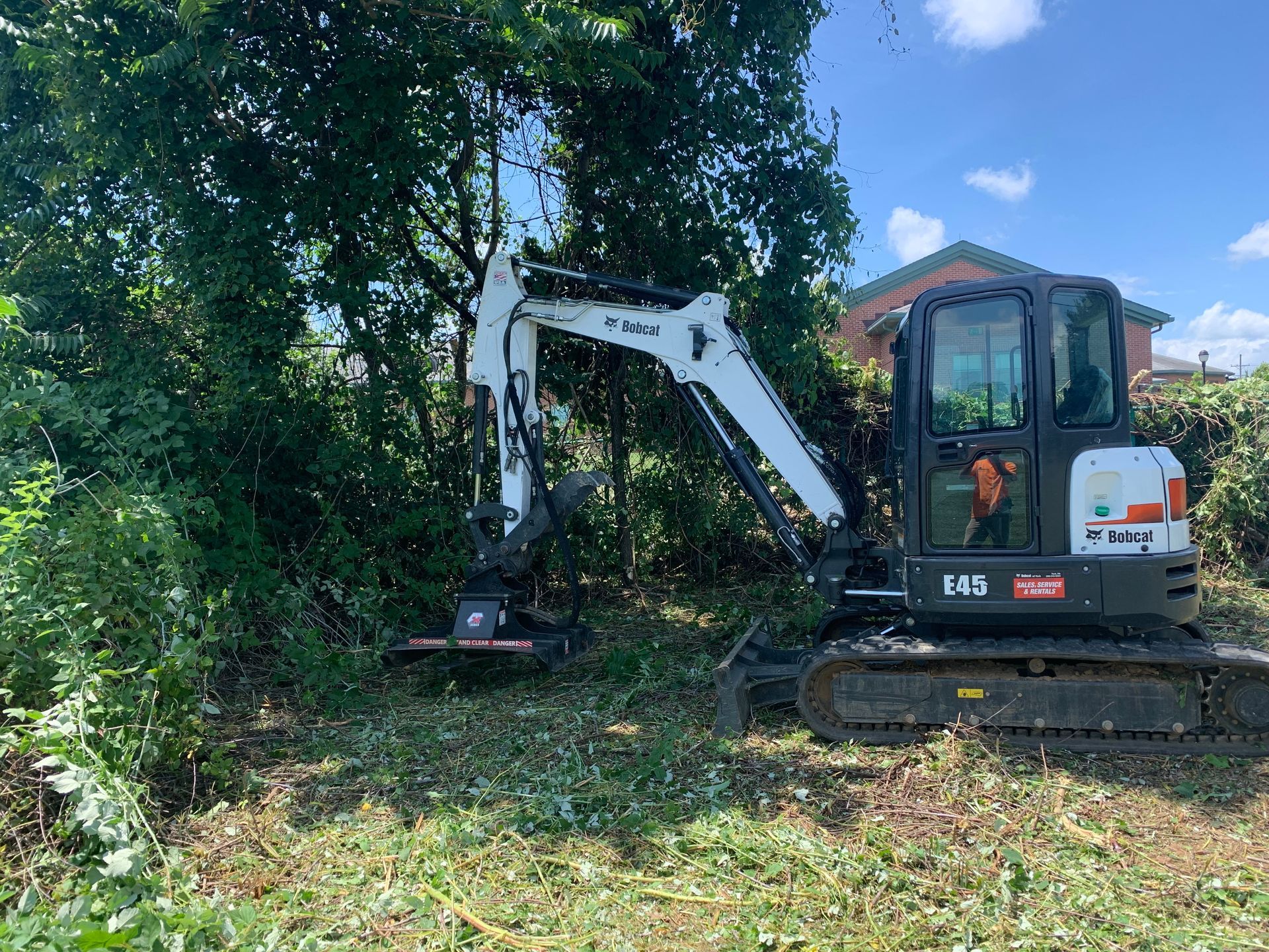 A white Bobcat excavator with a forestry mulcher attachment clearing brush in a sunny, wooded outdoor area.