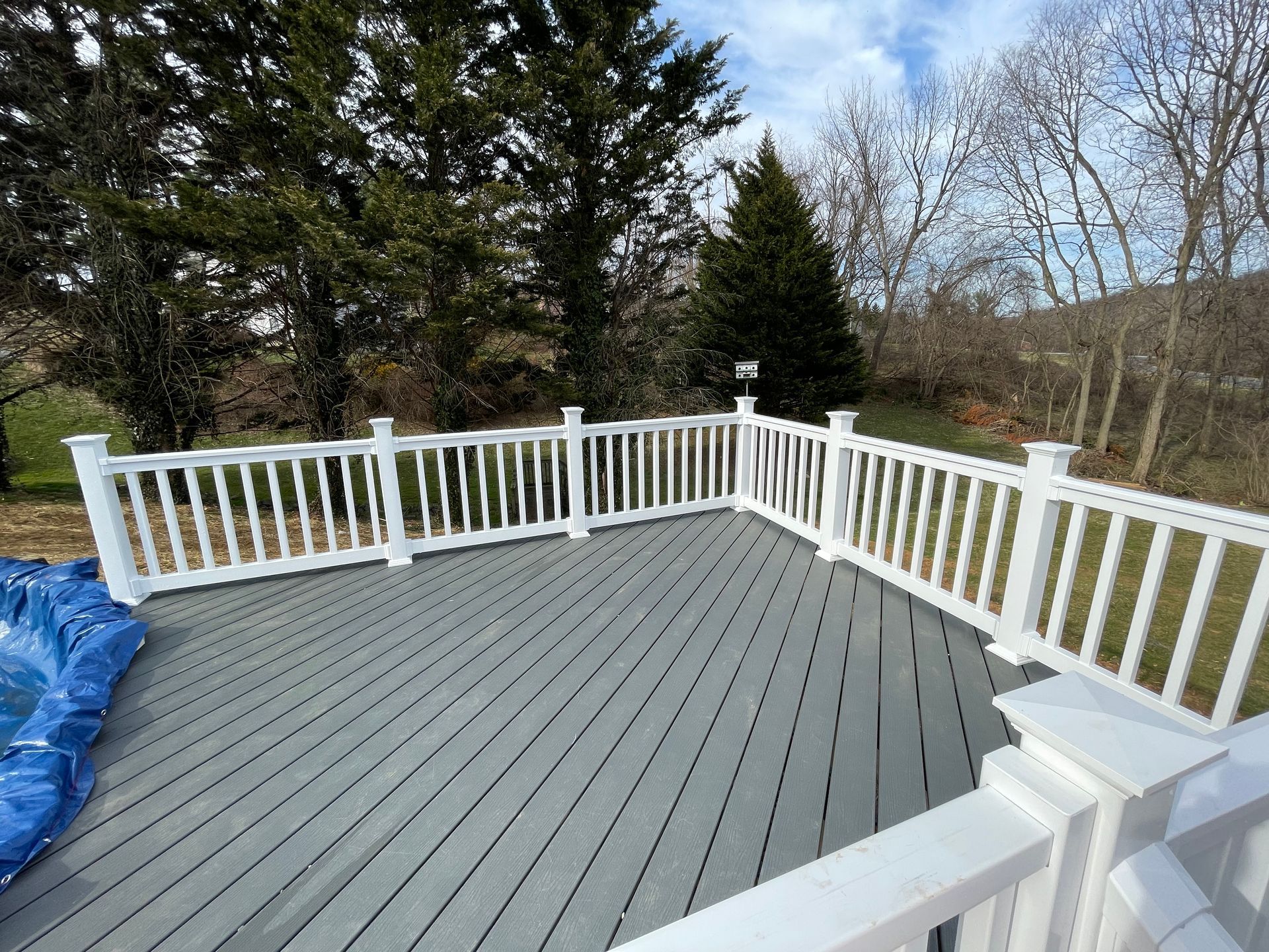 A gray deck with white railings overlooks a lawn, bare trees, and evergreen bushes under a partly cloudy sky.