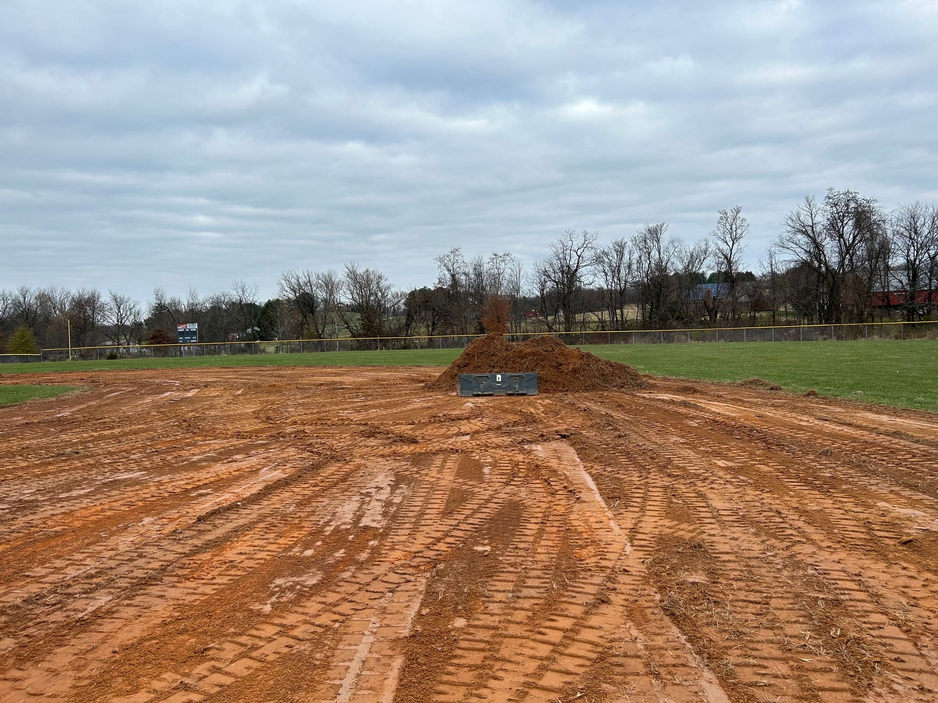 A construction site with tire tracks in reddish-brown soil, a mound of dirt, and a distant treeline under a cloudy sky.