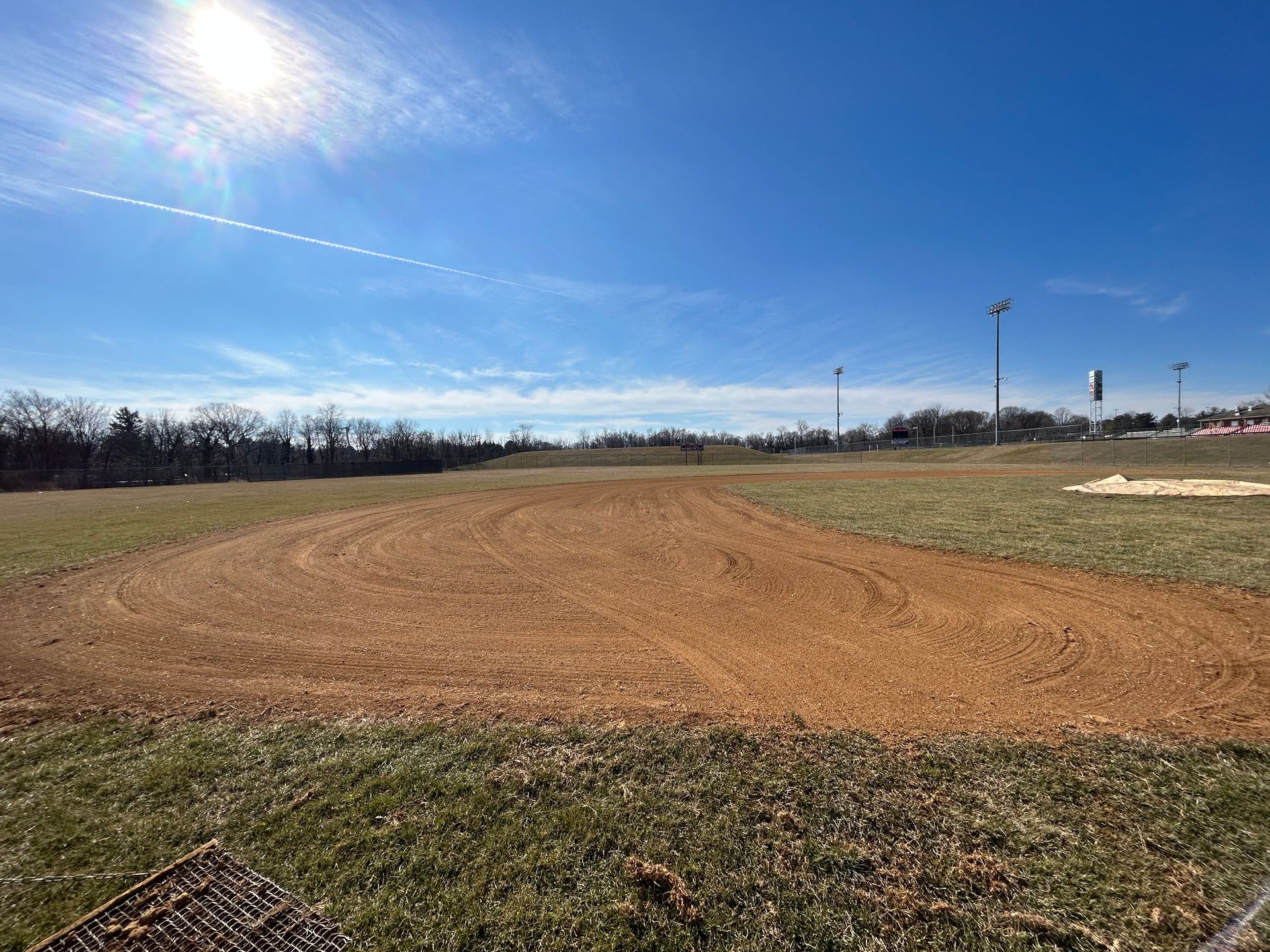 A dirt baseball infield sits under a bright blue sky with a visible sun flare and light clouds.