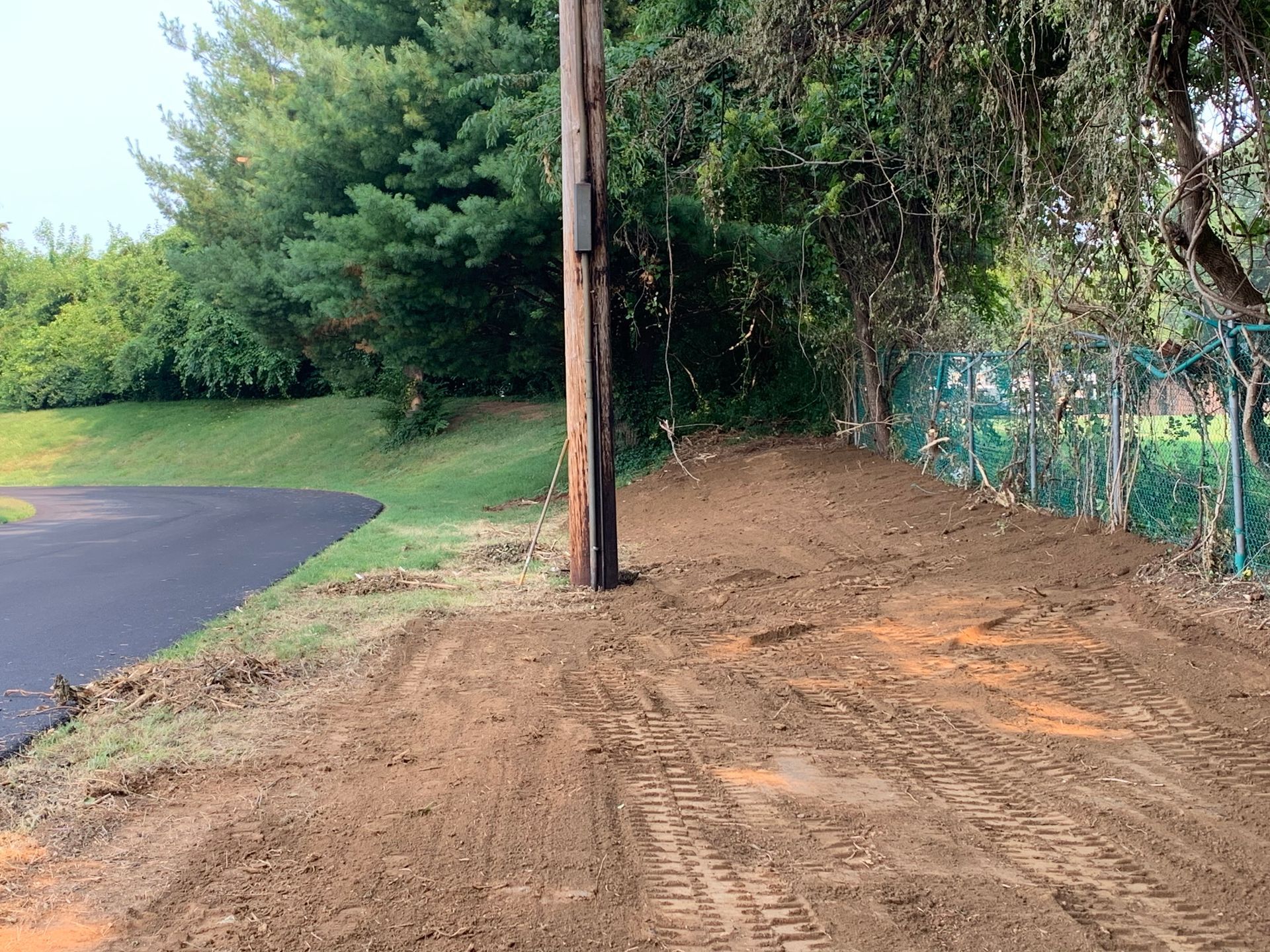 A wooden utility pole stands beside a freshly cleared dirt path next to a paved road and a chain-link fence.
