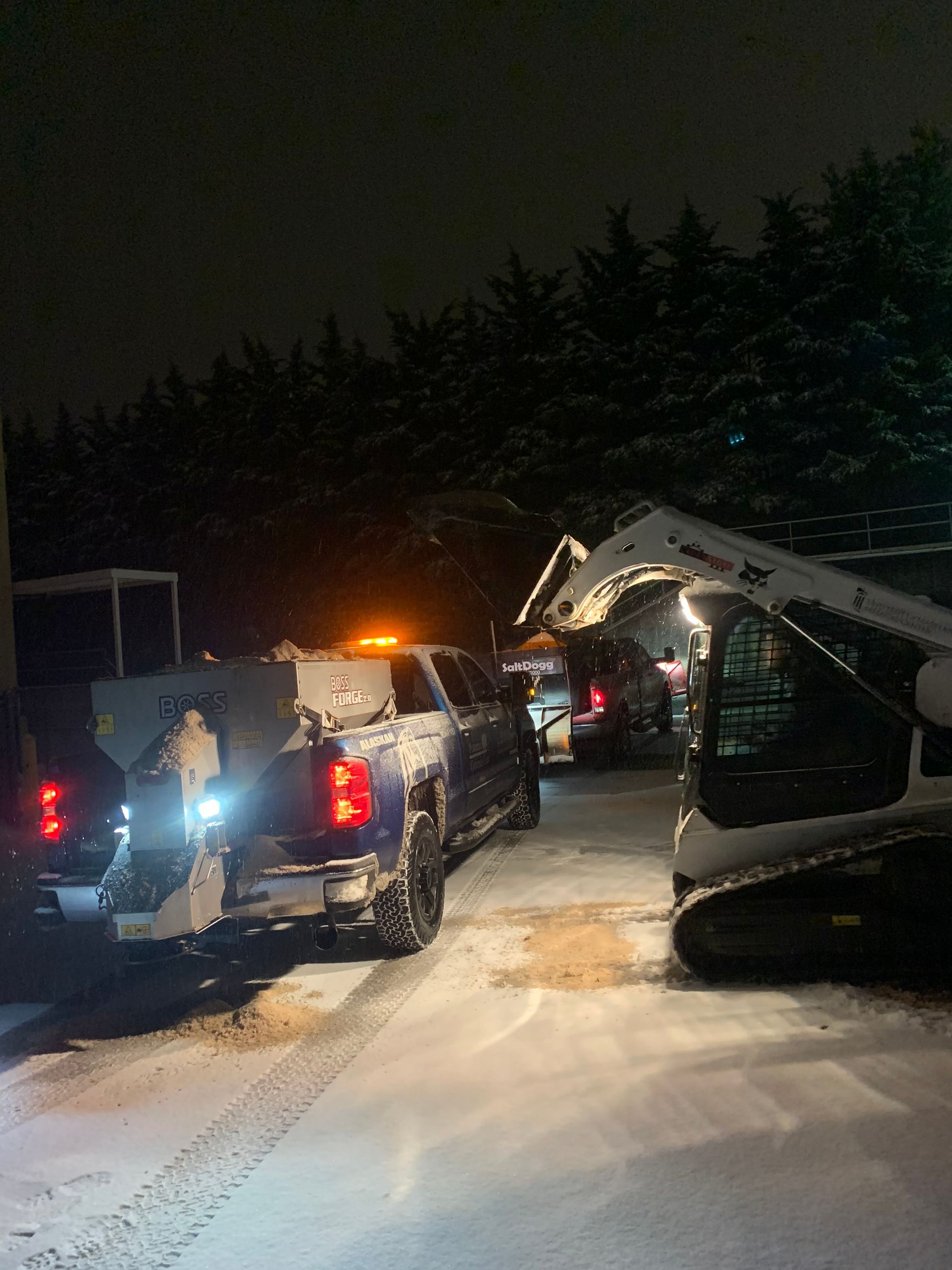 A truck and a skid-steer loader working on a snow-covered surface at night, with trees in the background.