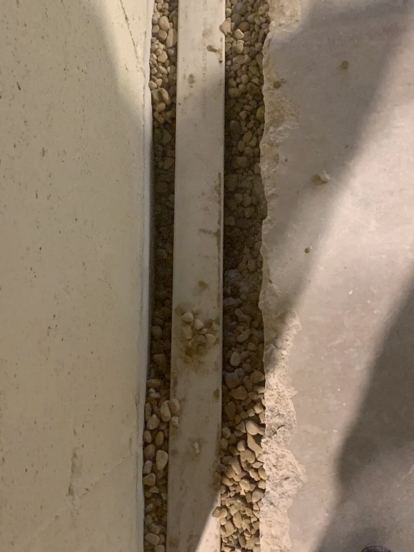 A section of a basement drainage system: gravel, a white pipe, and concrete walls and floor.