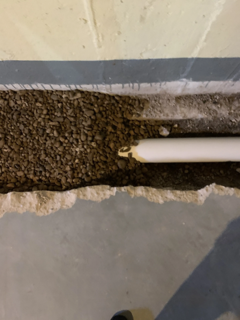Trench filled with gravel, containing a white PVC pipe, along the base of a concrete wall and floor.
