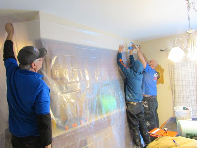 Three people install plastic sheeting on a wall and ceiling. One person holds the plastic, two apply tape. Interior setting.