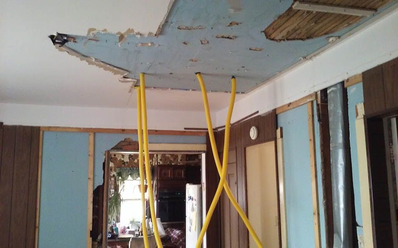 Damaged ceiling with exposed beams and yellow wires, room in disrepair, view into another room.