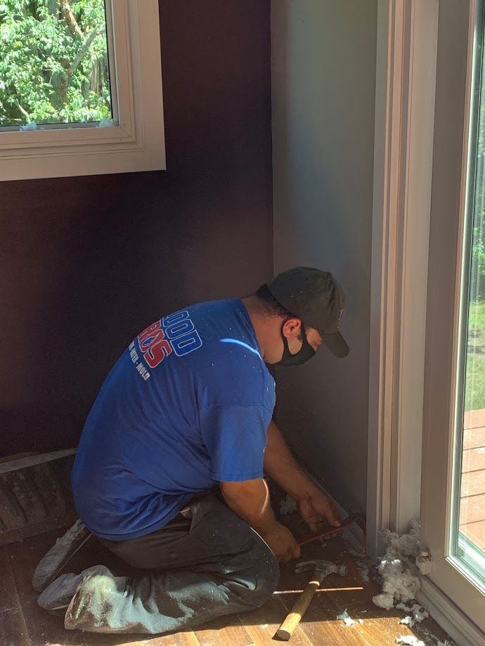 Person wearing a mask kneels, working on a floor near a window. Gray and purple walls, sunlight.