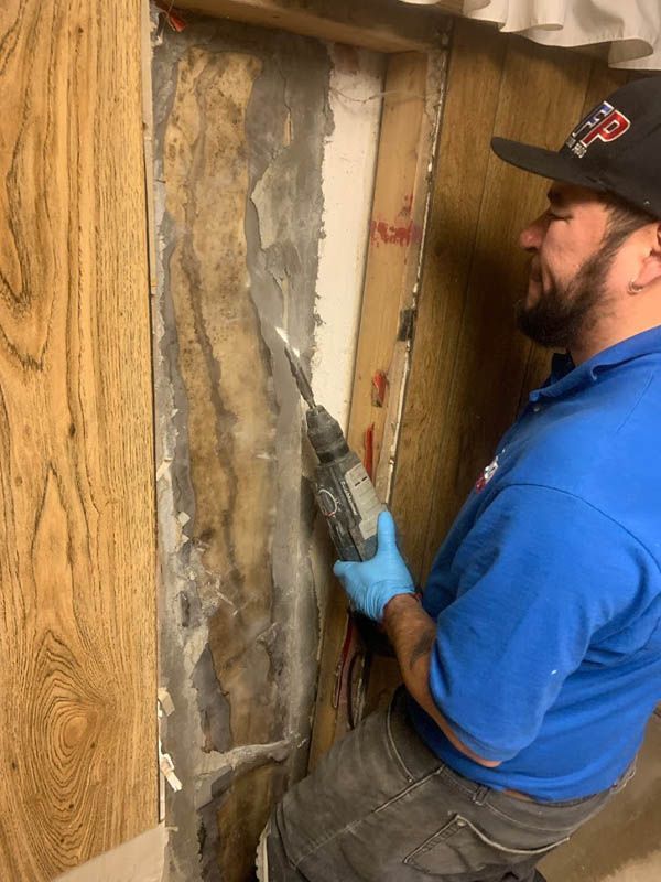Man in blue shirt using a drill on a wall with peeling material. Dark hair, beard, wearing a Patriots hat.