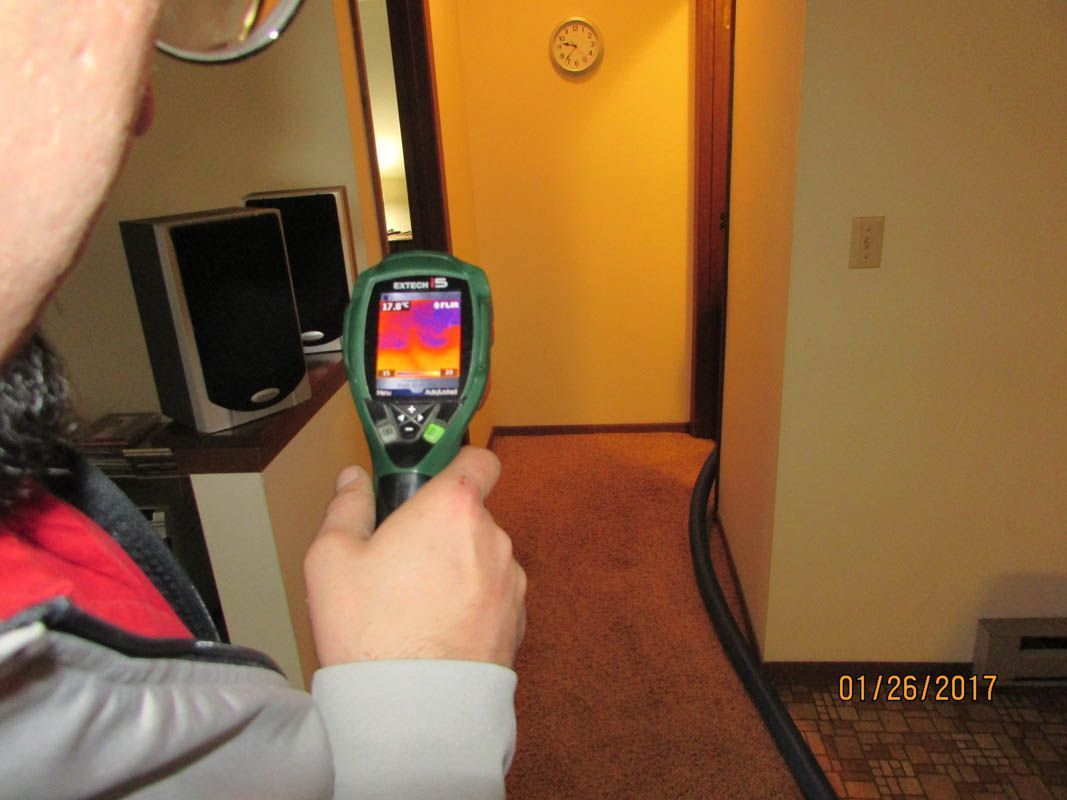 Person holding a thermal imaging camera in a hallway. The screen displays heat signatures, indicating temperature variations in the walls and floor.