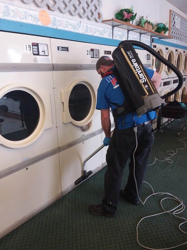 Man using a backpack vacuum to clean a laundromat floor near washing machines.