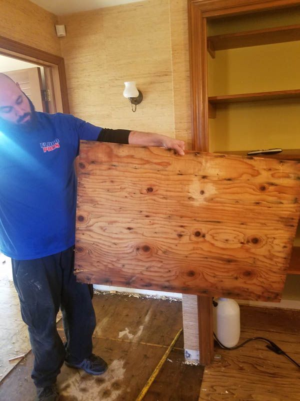 Man in blue shirt holds a large, stained wooden board indoors near a bookcase.